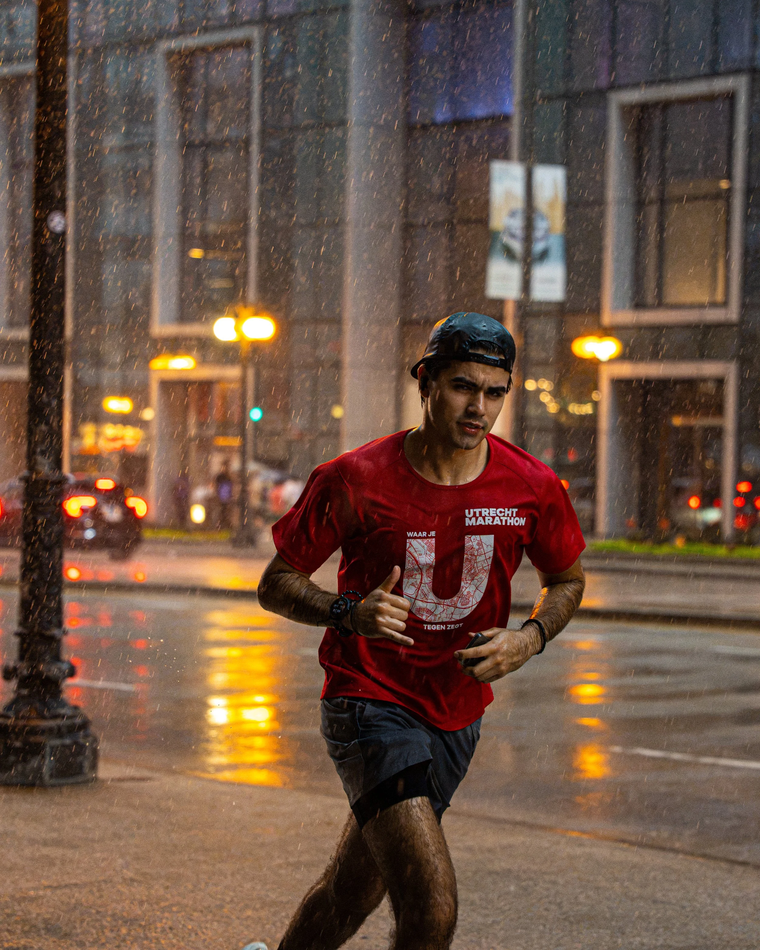 A man running in the rain at night on a city street, wearing a black cap and a red T-shirt with white graphics.