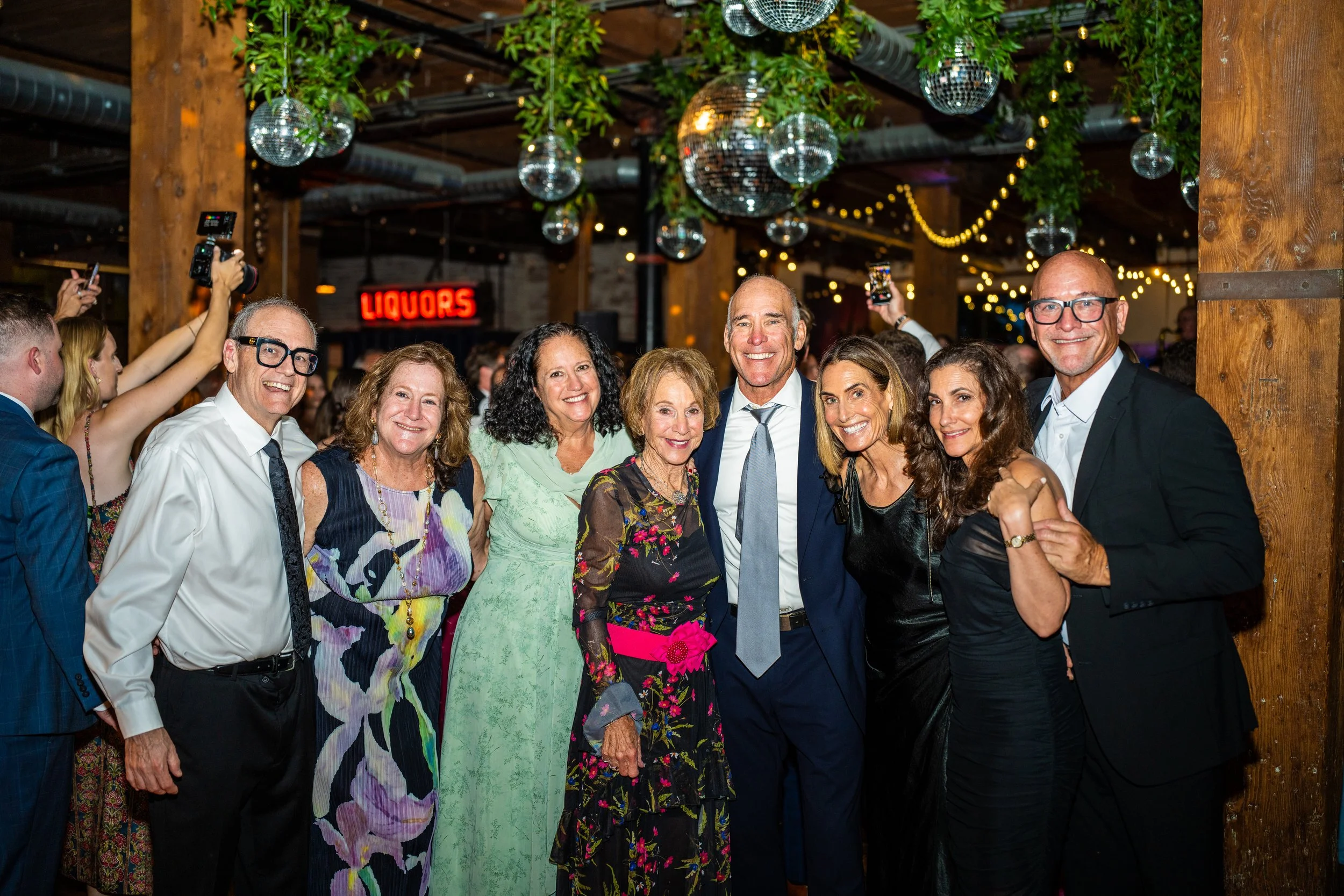 A group of people dressed in semi-formal attire smiling for a photo at a party or celebration, with a rustic wooden interior decorated with hanging disco balls and string lights, and a neon sign that reads 'LIQUORS' in the background.