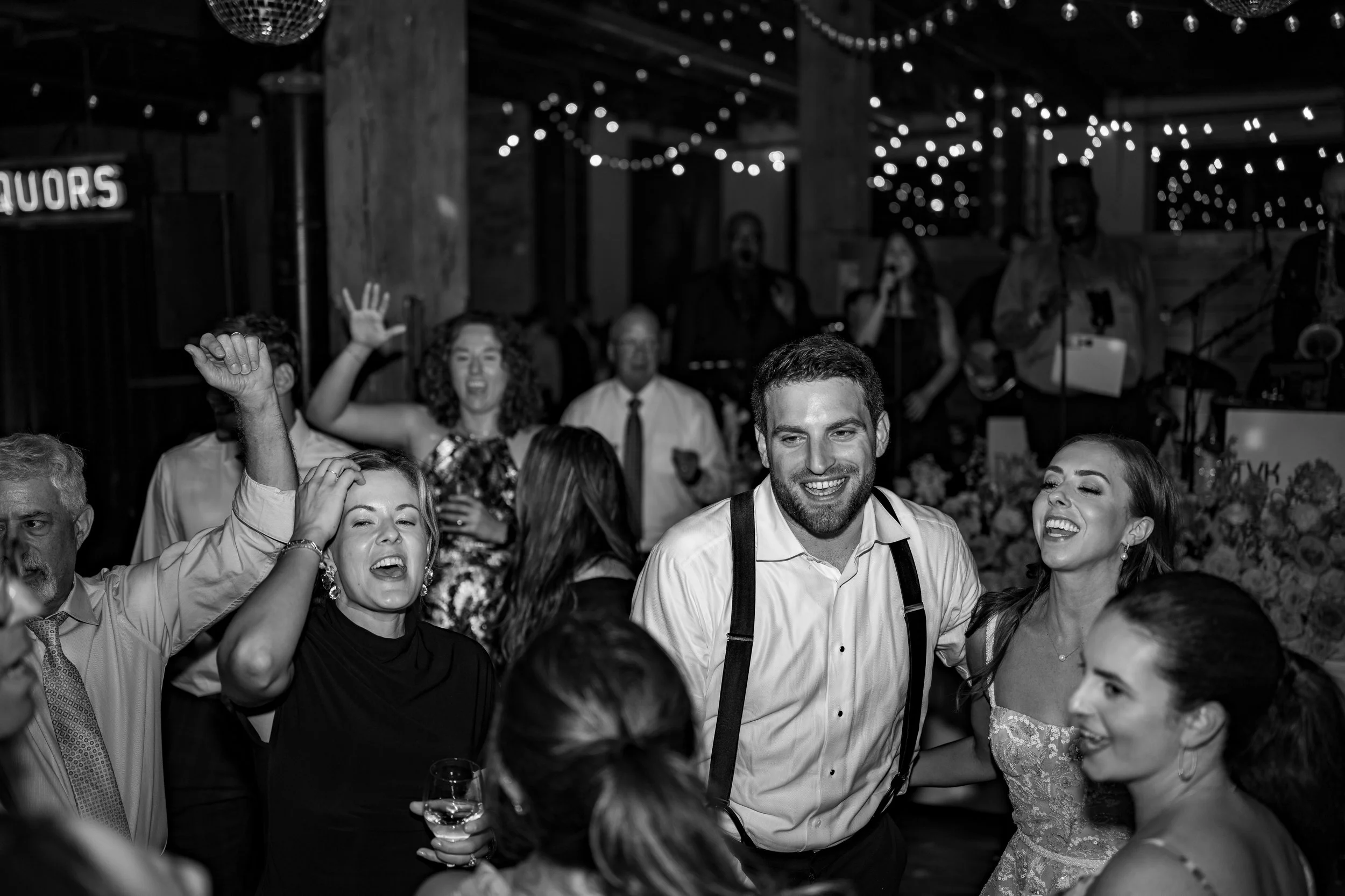 A black and white photo of a lively celebration with people dancing and smiling. In the background, musicians are performing on stage with string lights hanging overhead.