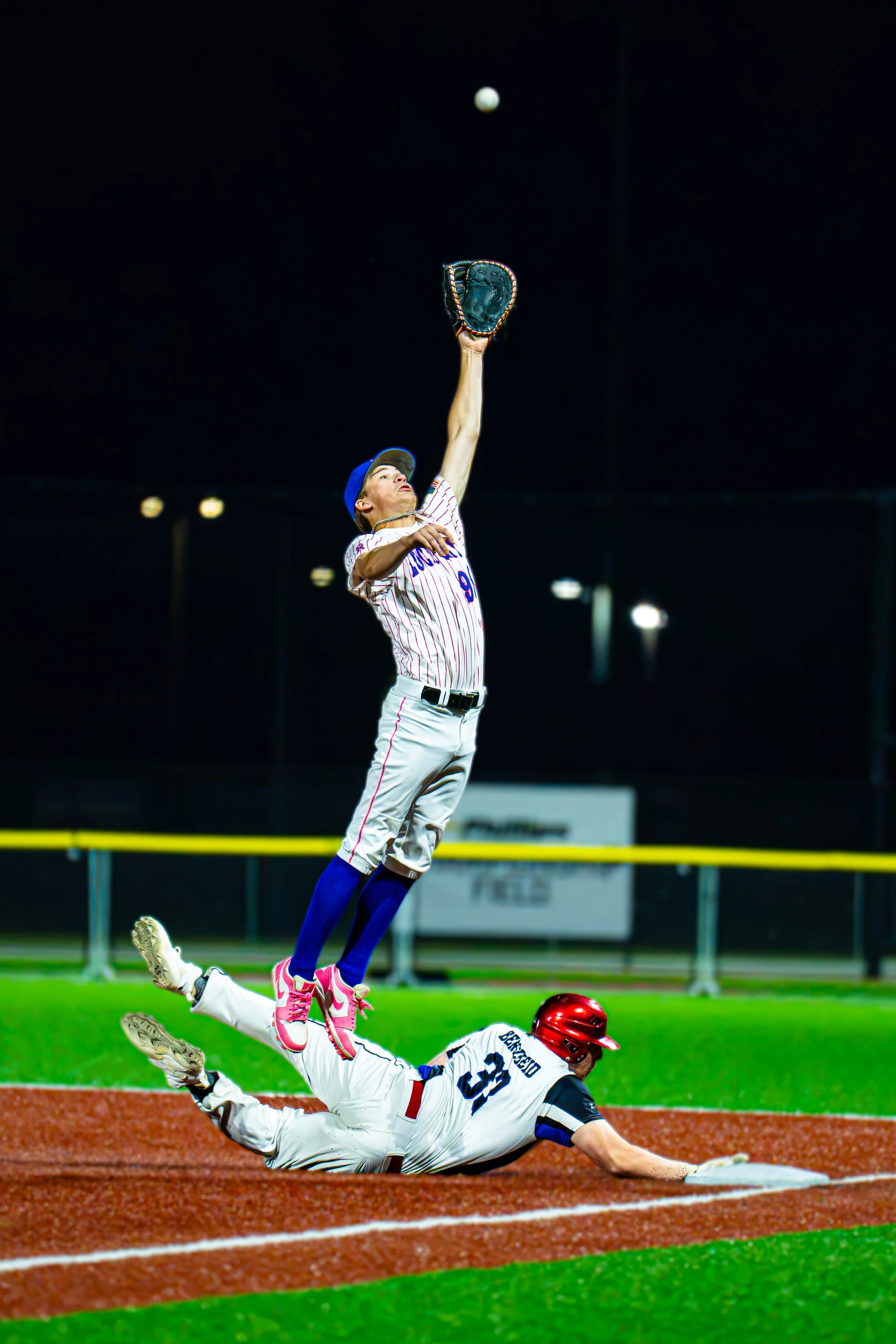 A baseball player in a blue and white uniform leaps to catch the ball at home plate while an opposing player, wearing a red and white uniform and helmet, slides into the base. The background is a baseball field with a scoreboard and lights.