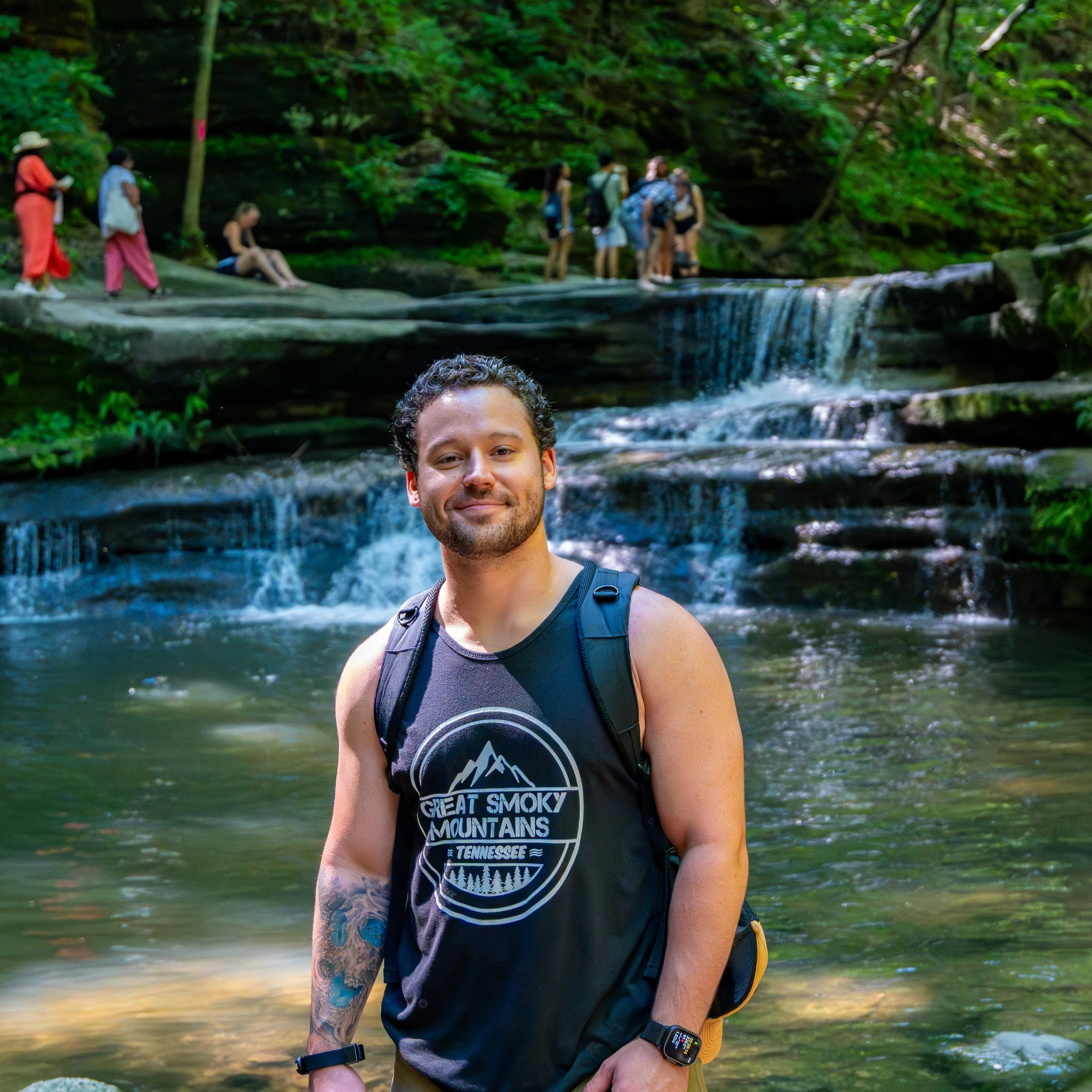 A young man with curly hair and tattoos on his right arm, smiling, standing in front of a small waterfall in a lush forest, wearing a black tank top with a mountain and trees graphic, a wristwatch, and a backpack.