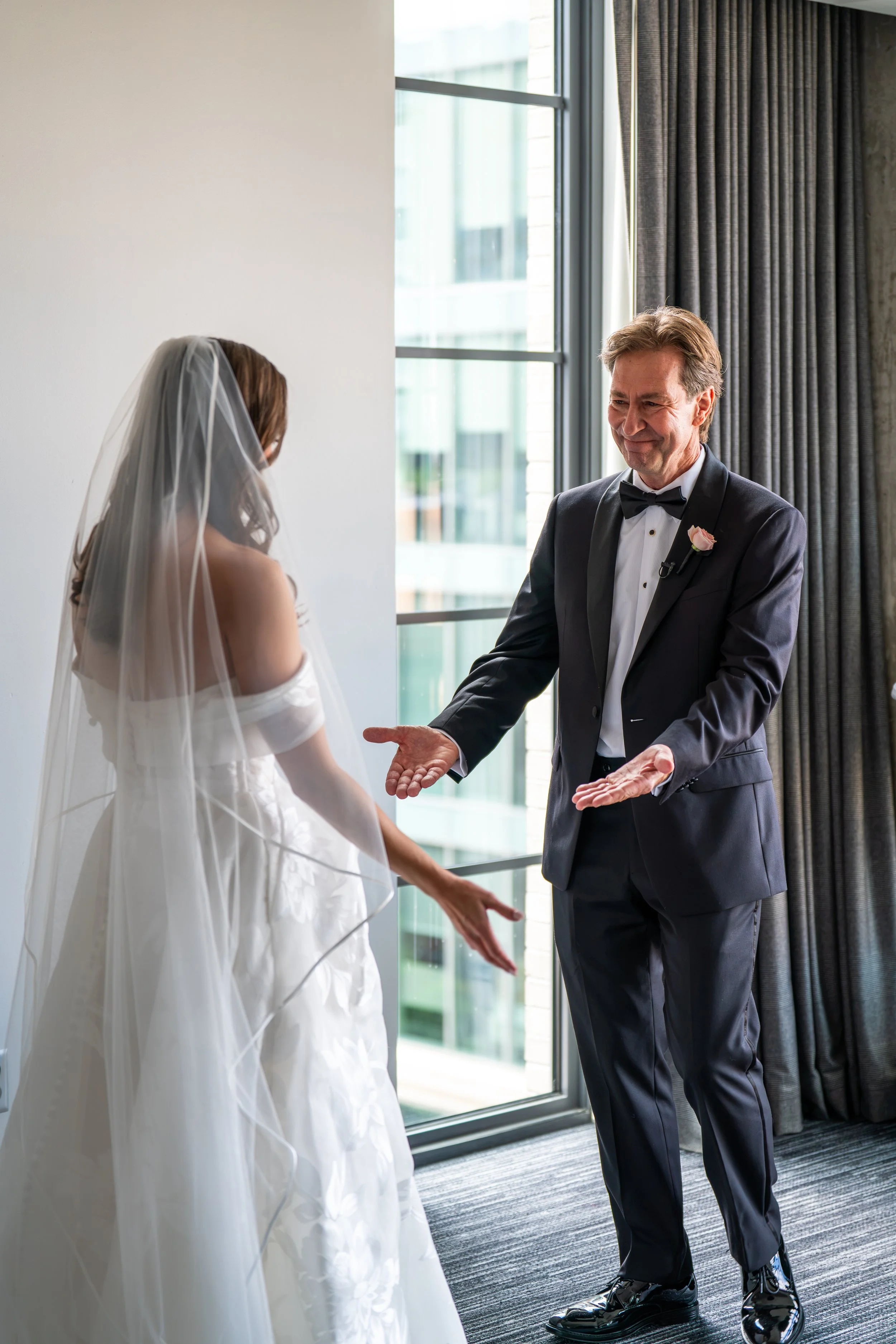A groom in a tuxedo with a pink boutonniere stands with his hands extended, smiling, during a wedding ceremony with a bride in a white wedding dress and veil, inside a modern room with large windows and gray curtains.