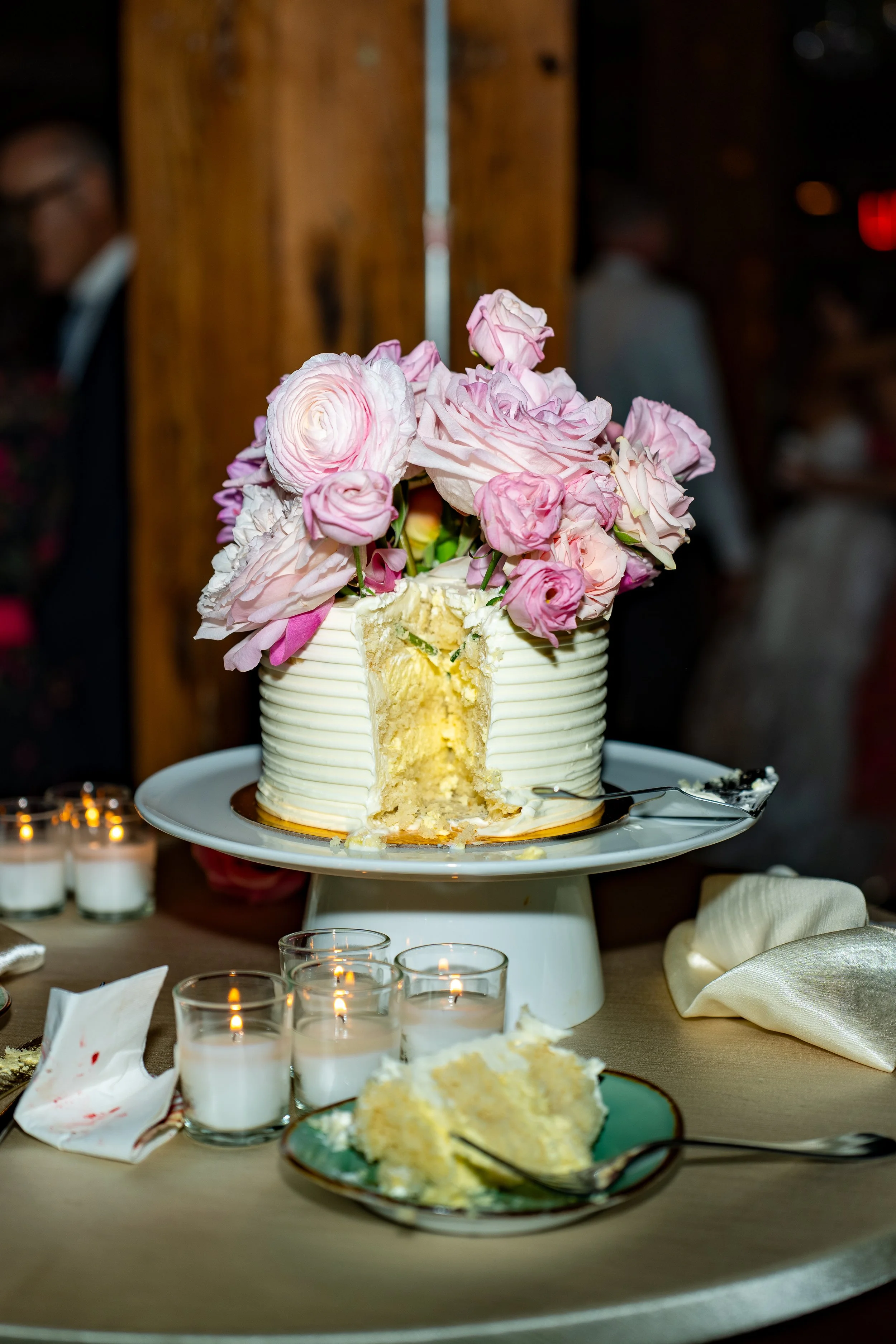 A partially cut wedding cake topped with pink and white roses on a cake stand, surrounded by candles and a plate with cake on a table.