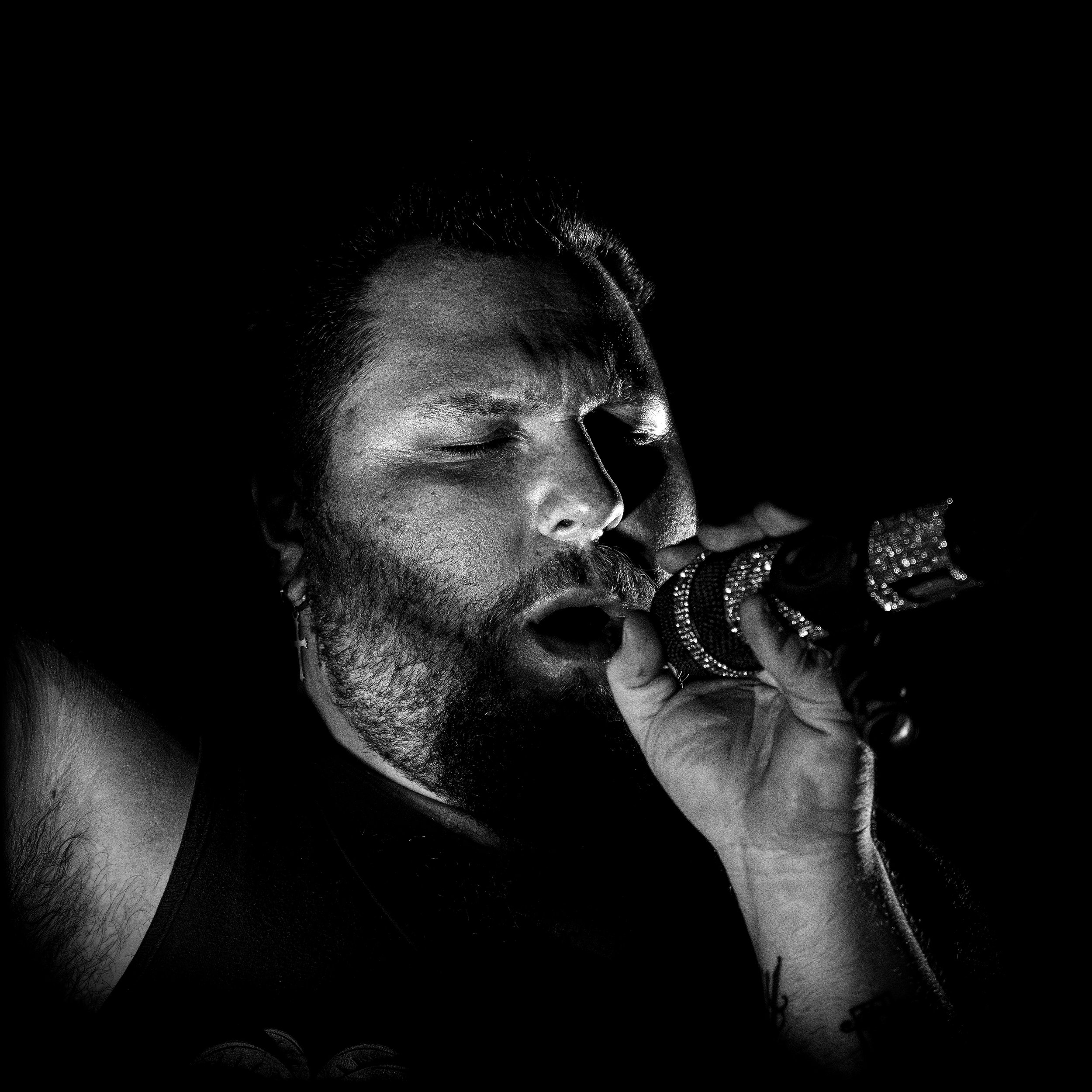 A person with a beard and closed eyes holding a microphone close to their face, singing or speaking with their face slightly illuminated against a dark background.