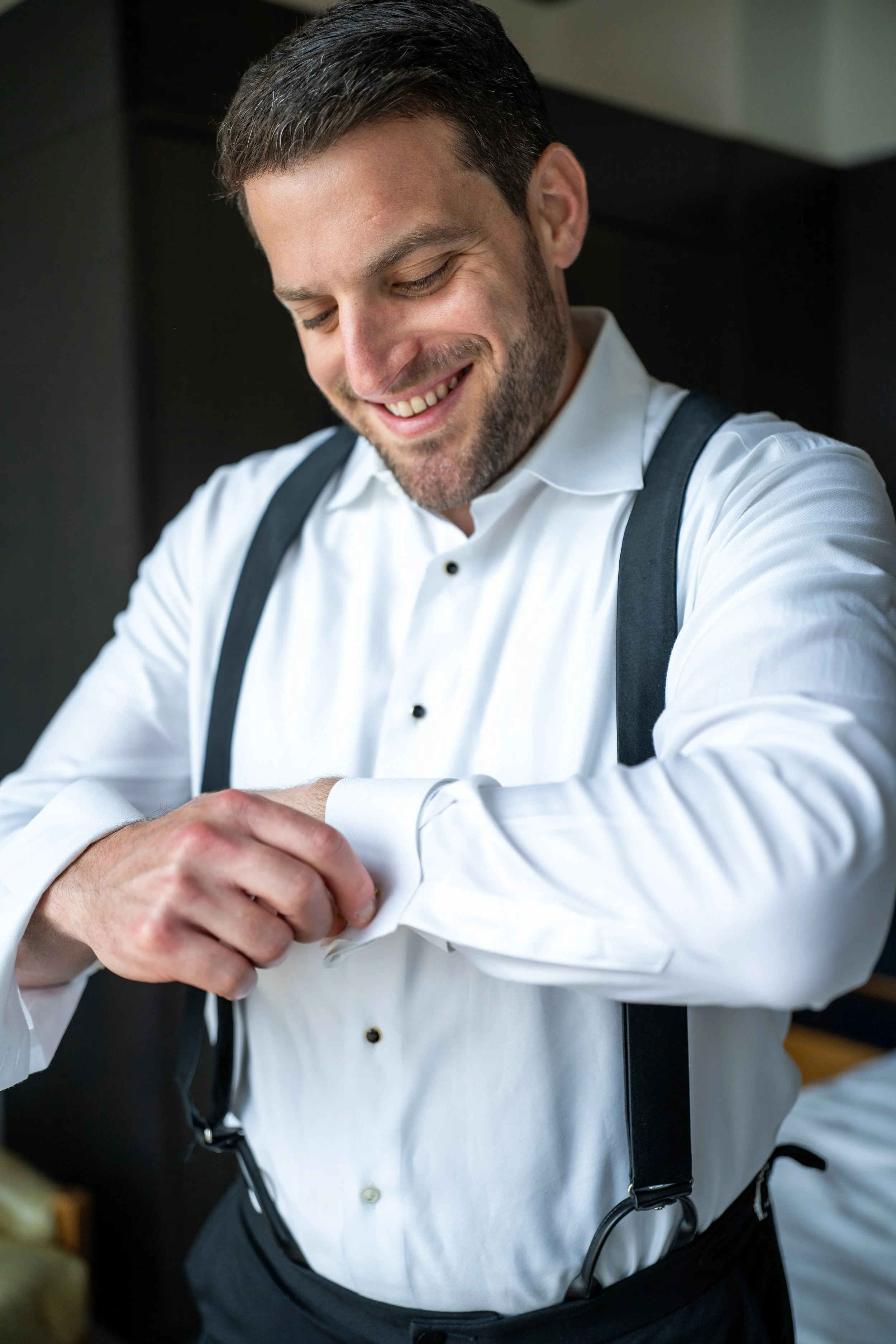 Smiling man with dark hair and beard adjusting his white dress shirt cuff, wearing suspenders, standing in a room with dark background.