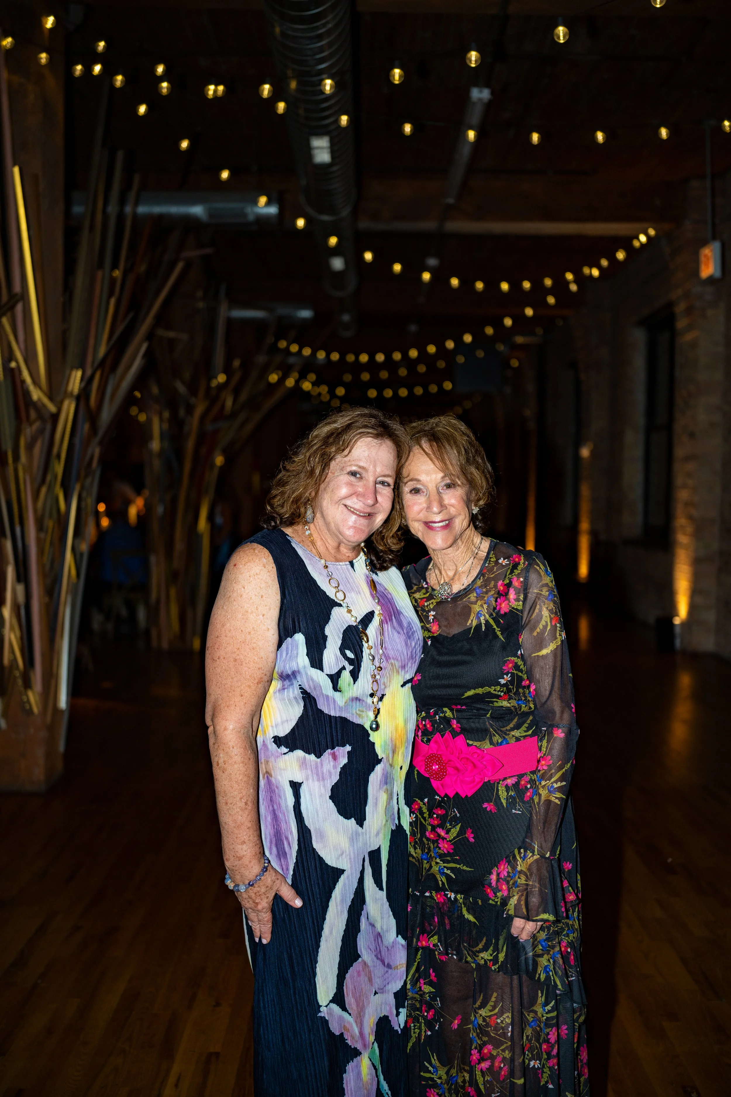 Two women smiling and standing close together at an indoor event, with wooden floors, brick walls, and string lights in the background.