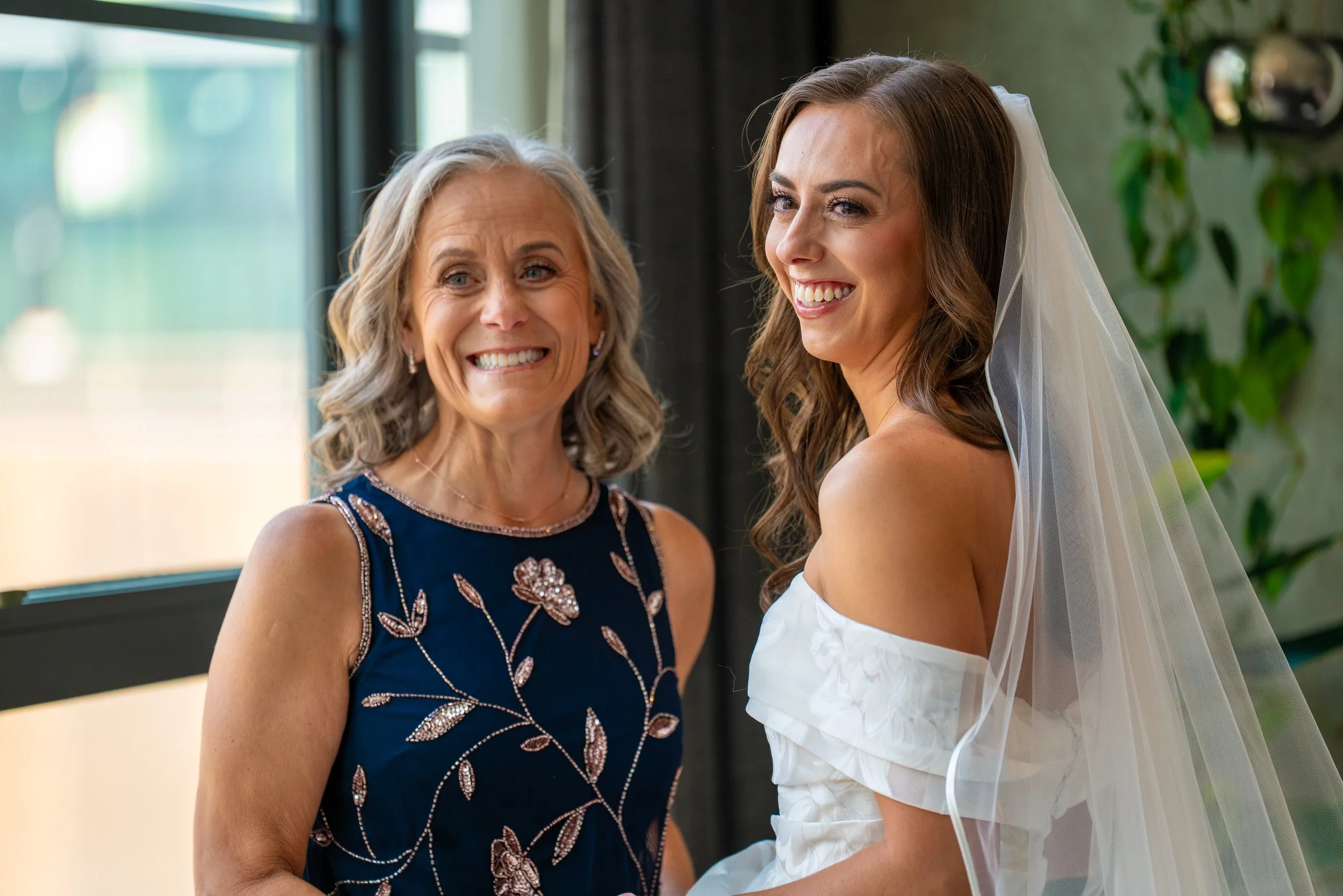 A bride with long brown hair and a veil smiling with an older woman, both standing by a window with greenery outside.