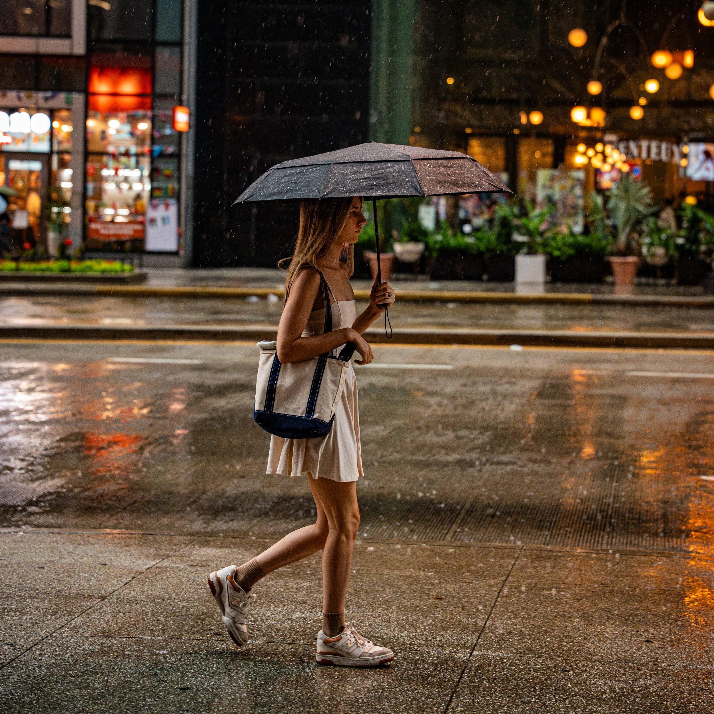 A woman walking in the rain at night with an umbrella, wearing a beige sundress, white sneakers, and carrying a tote bag, with blurred store lights in the background.