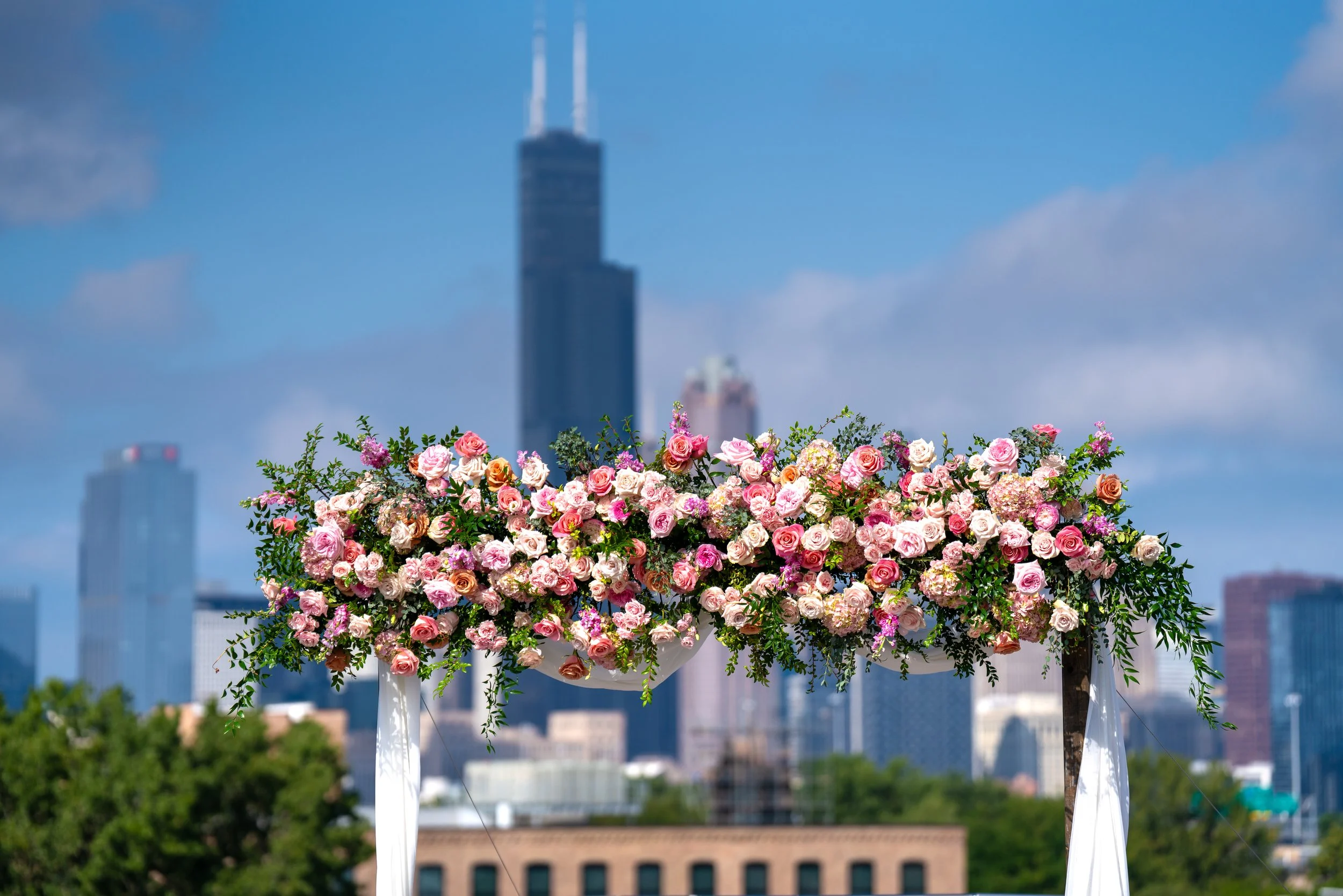 A large arrangement of pink, peach, and white roses and green foliage on a rooftop with a city skyline, including the One World Trade Center, in the background under a partly cloudy blue sky.