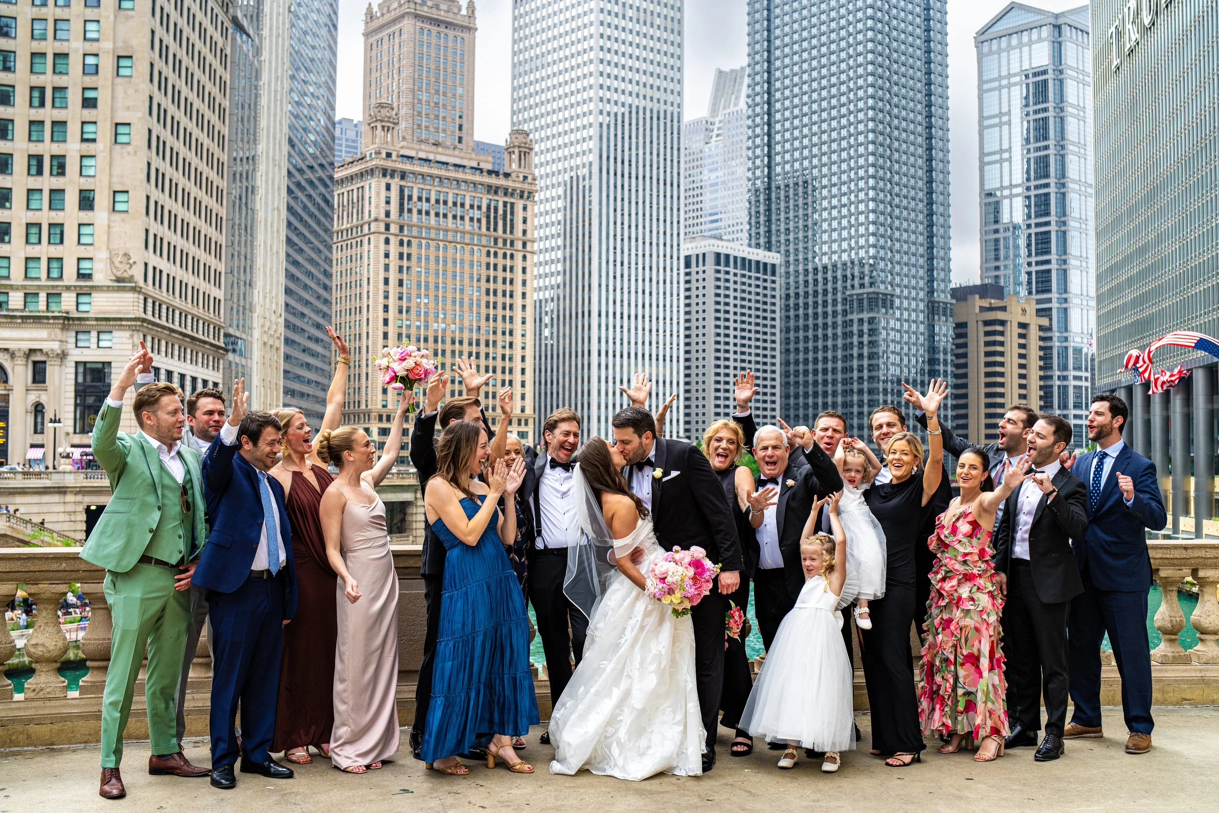 A large group of people celebrating on a bridge in an urban area with tall skyscrapers in the background, including a bride and groom at the center.