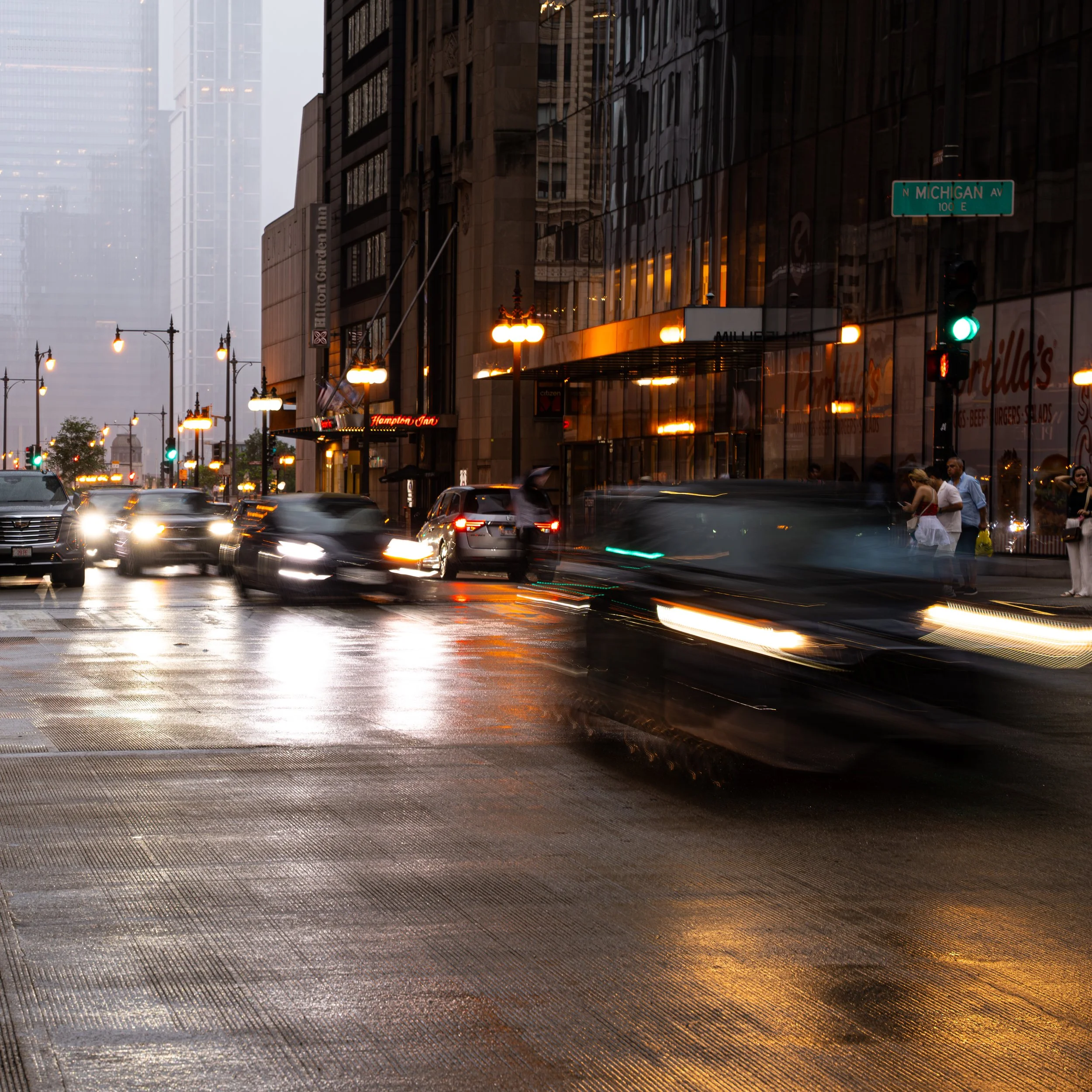 A city street scene at dusk with blurred moving cars, illuminated streetlights, and tall buildings, some with reflective glass windows, visible in the background.
