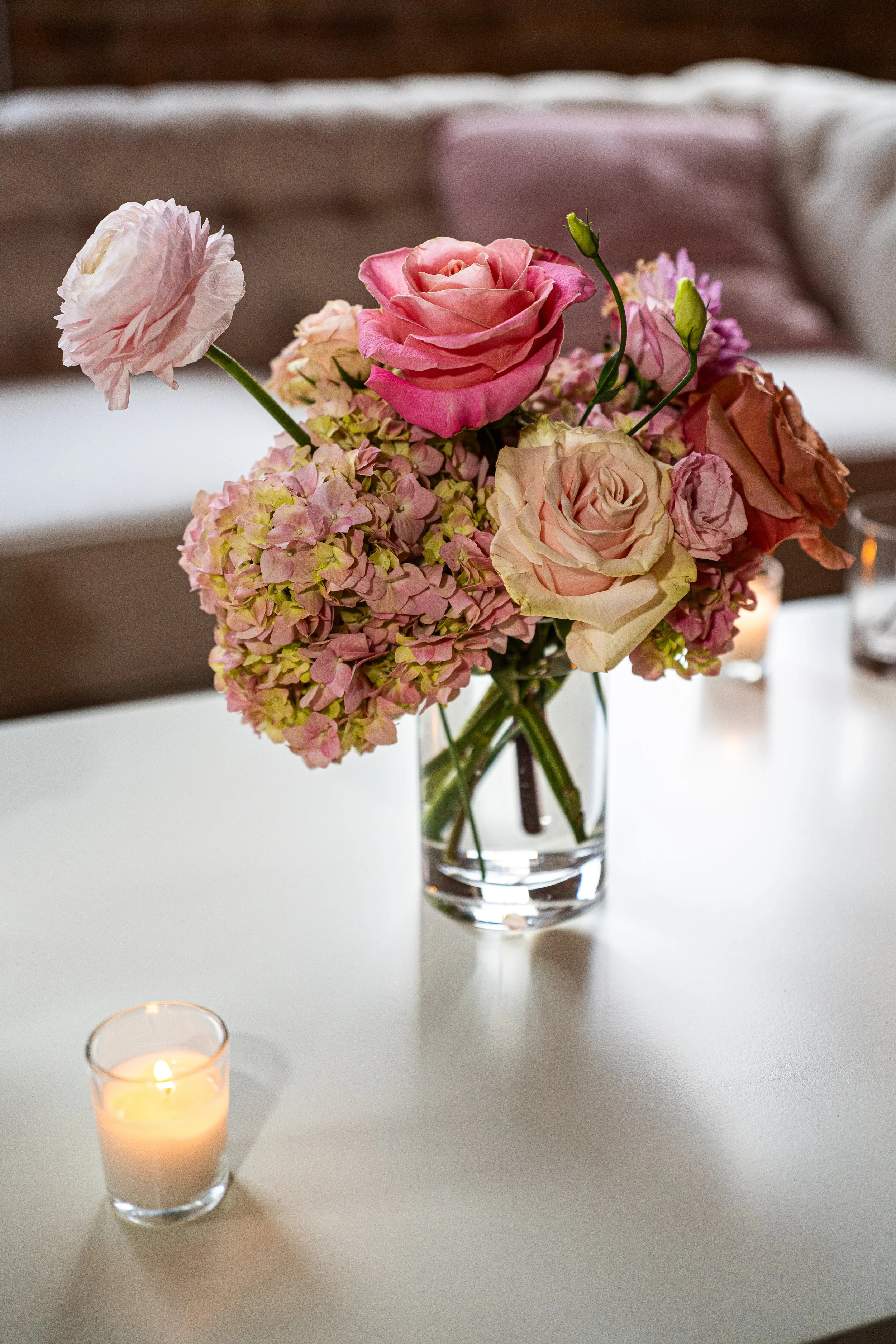 A bouquet of pink and white roses and hydrangeas in a clear glass vase on a white table, with a lit candle in a glass holder nearby.