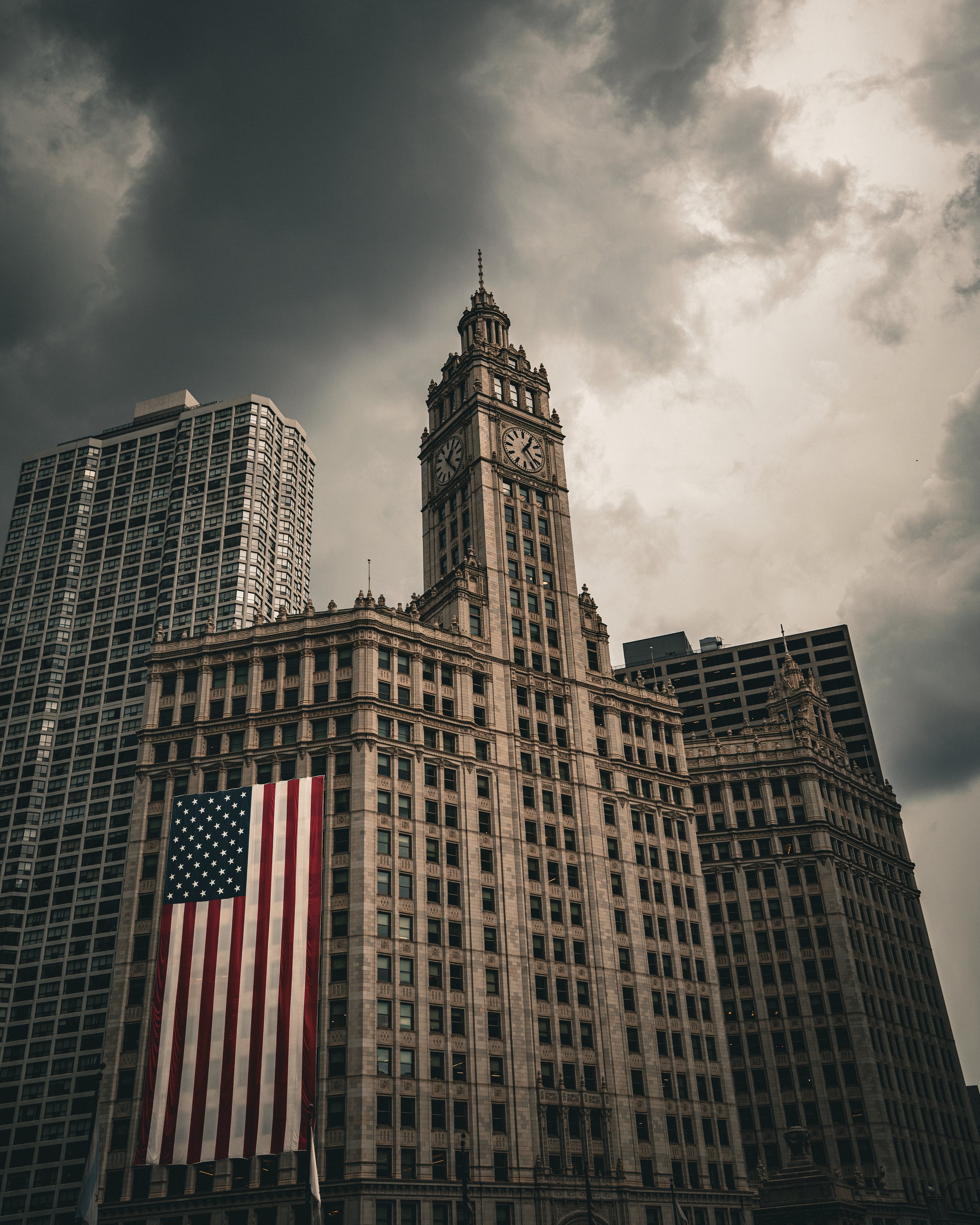 A large American flag hanging vertically on the side of a historic clock tower building in a cityscape under a cloudy sky.