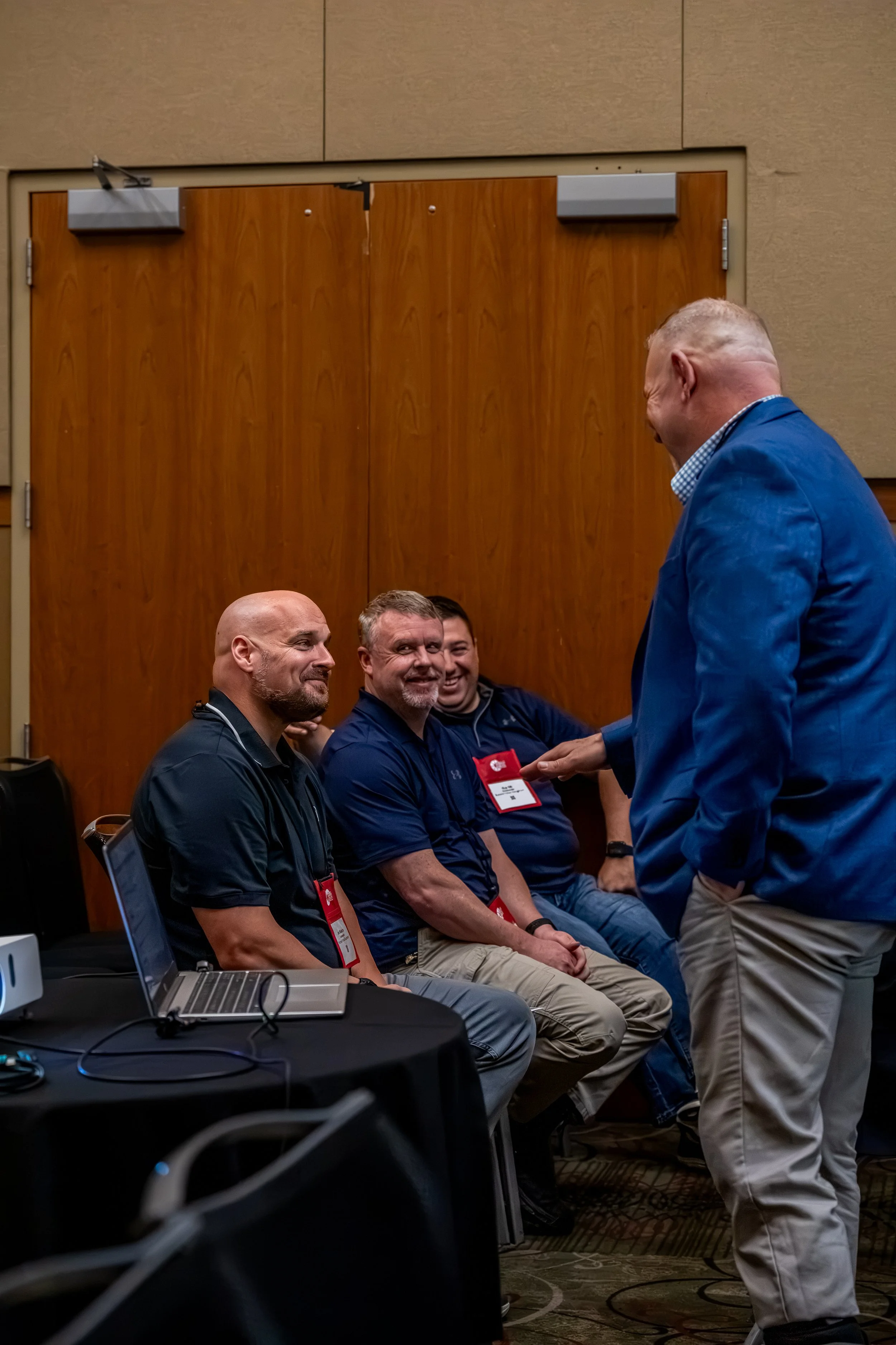 A group of men in a conference room engaged in conversation, some of them seated and one standing, with a laptop and conference badges visible.