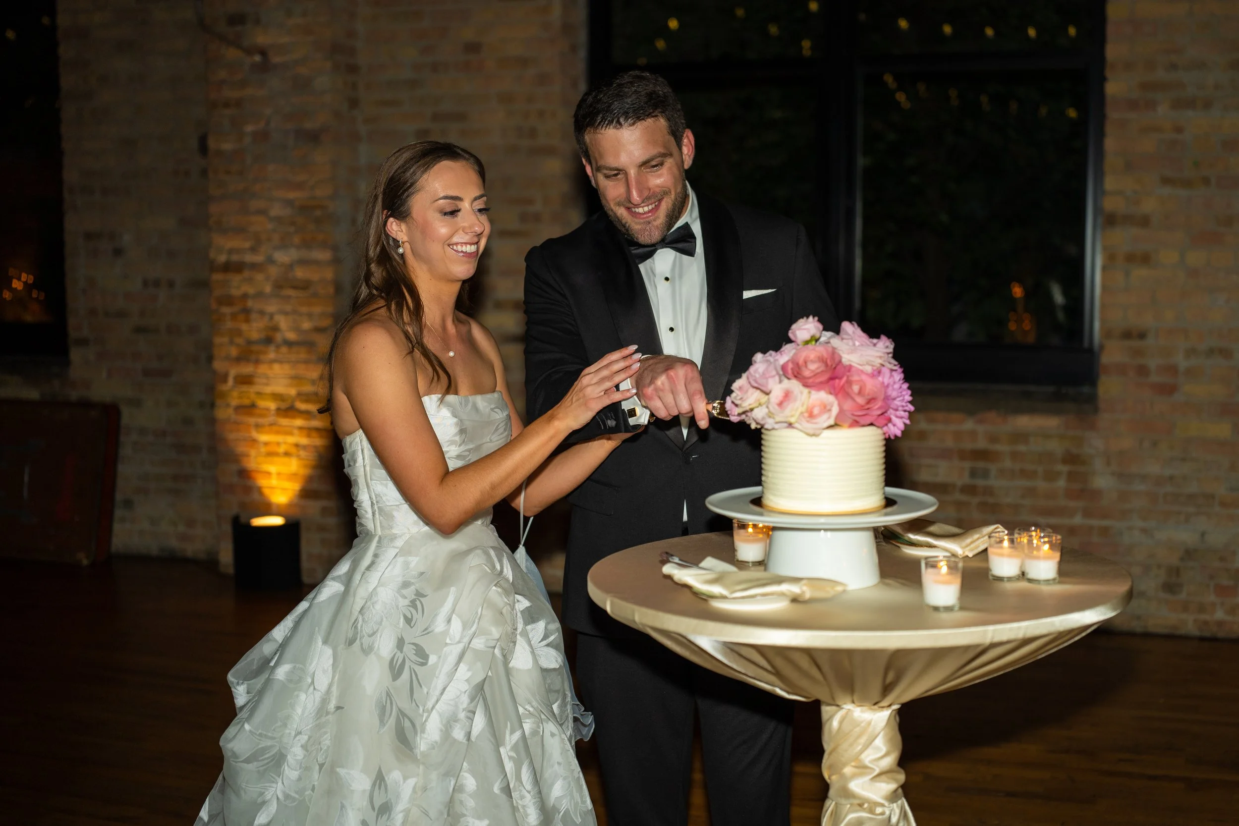 A bride and groom at a wedding, cutting a wedding cake together, smiling, in a dimly lit venue with brick walls and candles.