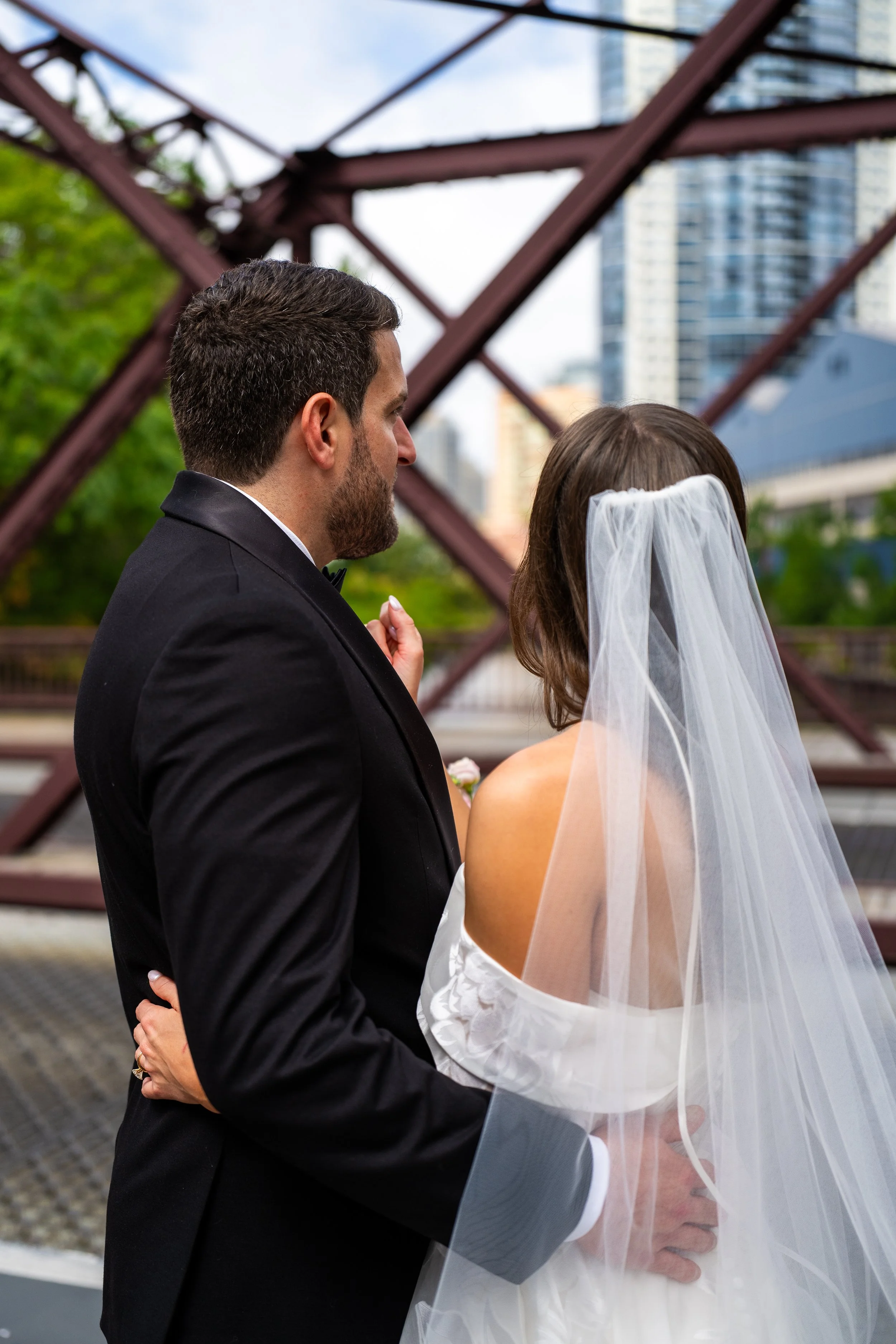 A bride and groom embrace outdoors in front of a rusty metal structure with city buildings in the background, the groom in a black tuxedo and the bride in a white dress with a sheer veil.