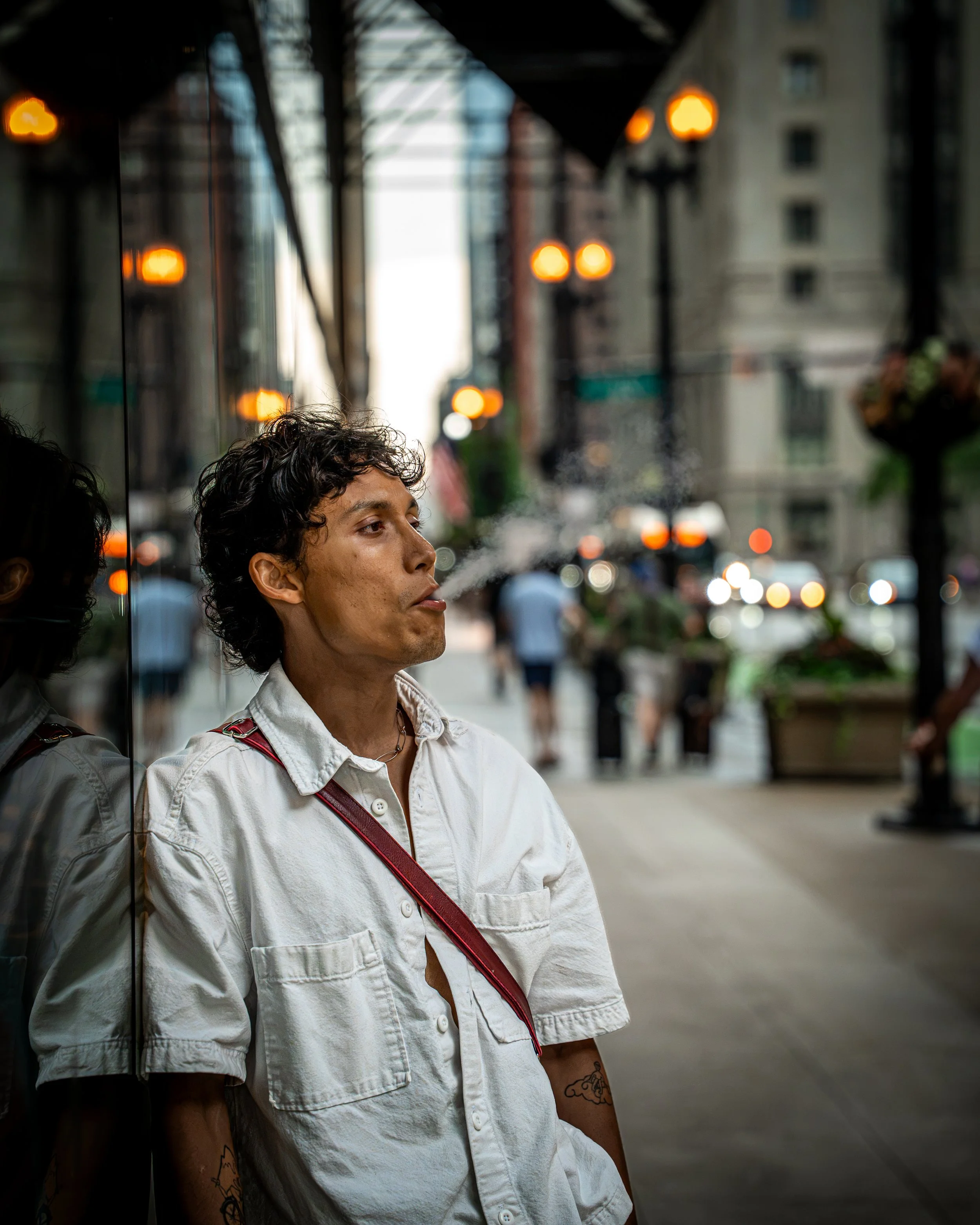 A young man with curly dark hair and tattoos on his arm, wearing a white jacket and red strap, is standing against a glass wall on a city street during dusk, with blurred city lights and buildings in the background.