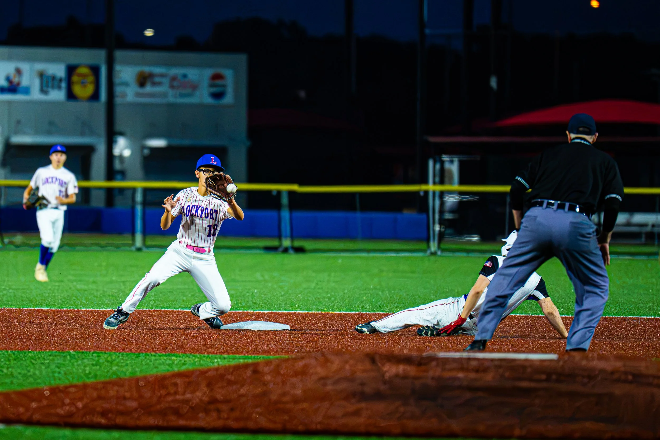 A youth baseball game during evening with players in uniform, one sliding into a base, another preparing to catch, and an umpire watching, on a field with green grass and dirt base paths.