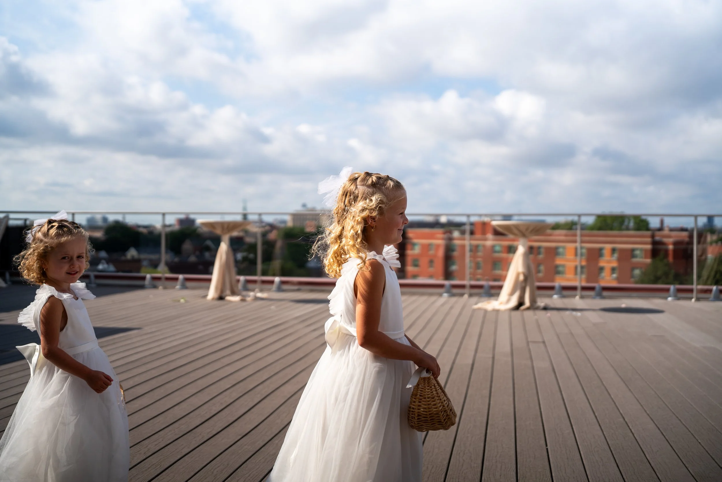 Two young girls with curly blonde hair wearing white dresses with bows, standing on a rooftop with city buildings in the background and a cloudy sky.