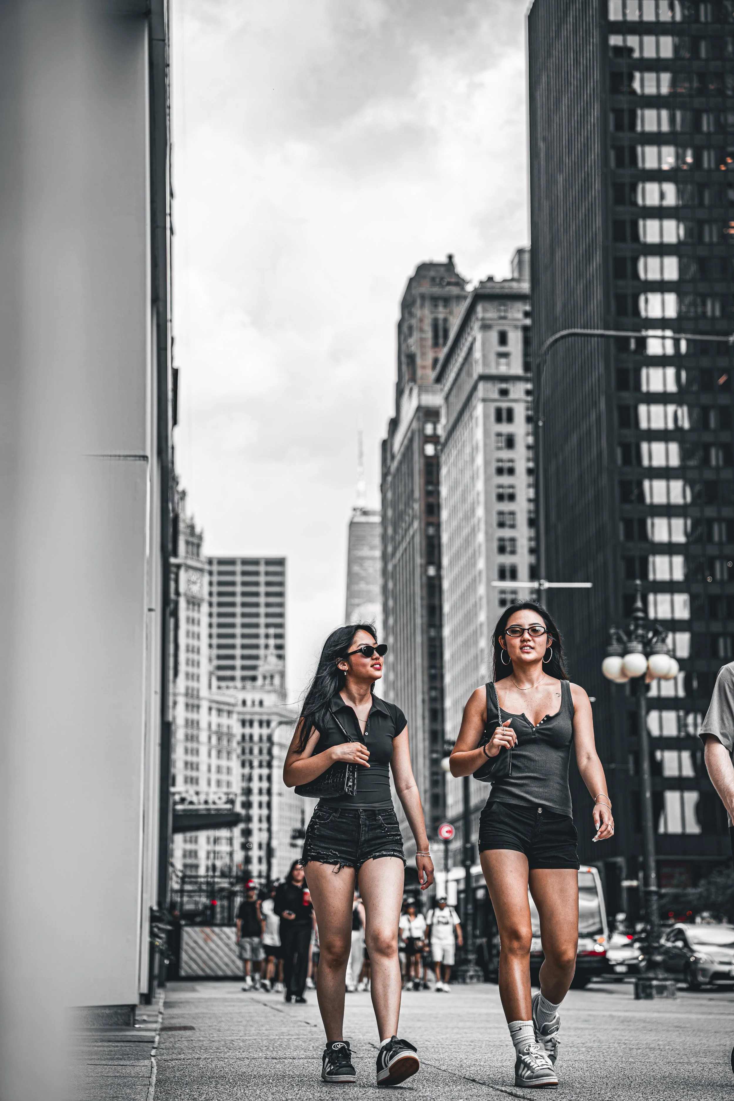 Two women walking down a city street with tall buildings in the background. One woman has long black hair, glasses, and wears a black top and shorts. The other has long black hair, glasses, and wears a black tank top and shorts. Both are wearing snea