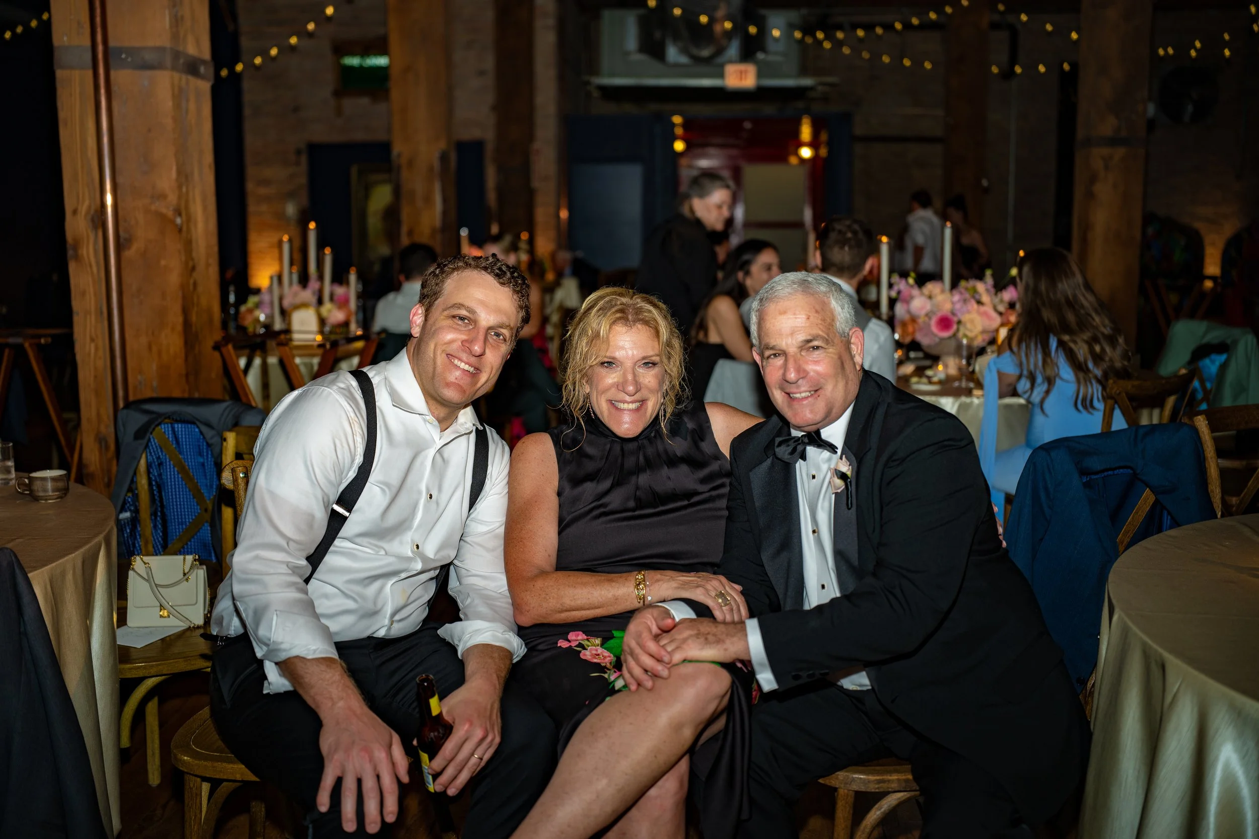 Three people sitting together at a formal event, smiling at the camera. They are dressed in formal attire, with the man on the left in a white shirt and suspenders, the woman in the middle in a black dress, and the man on the right in a tuxedo. The b