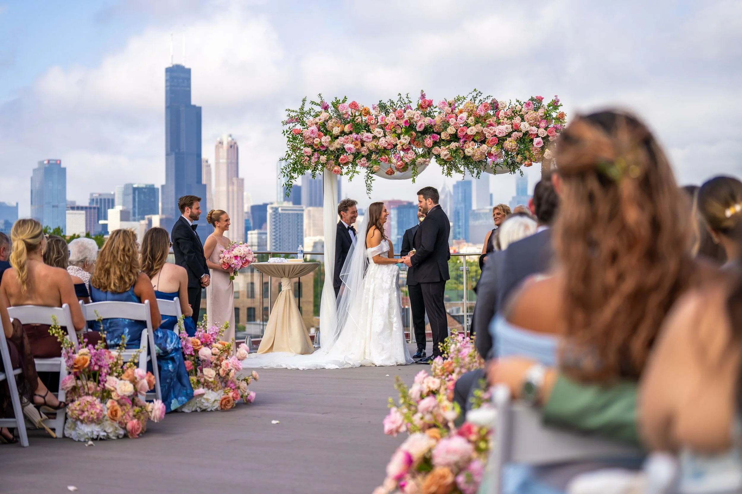 A wedding ceremony on a rooftop with the skyline of a city in the background, decorated with pink and white flowers, and a bride and groom exchanging vows.