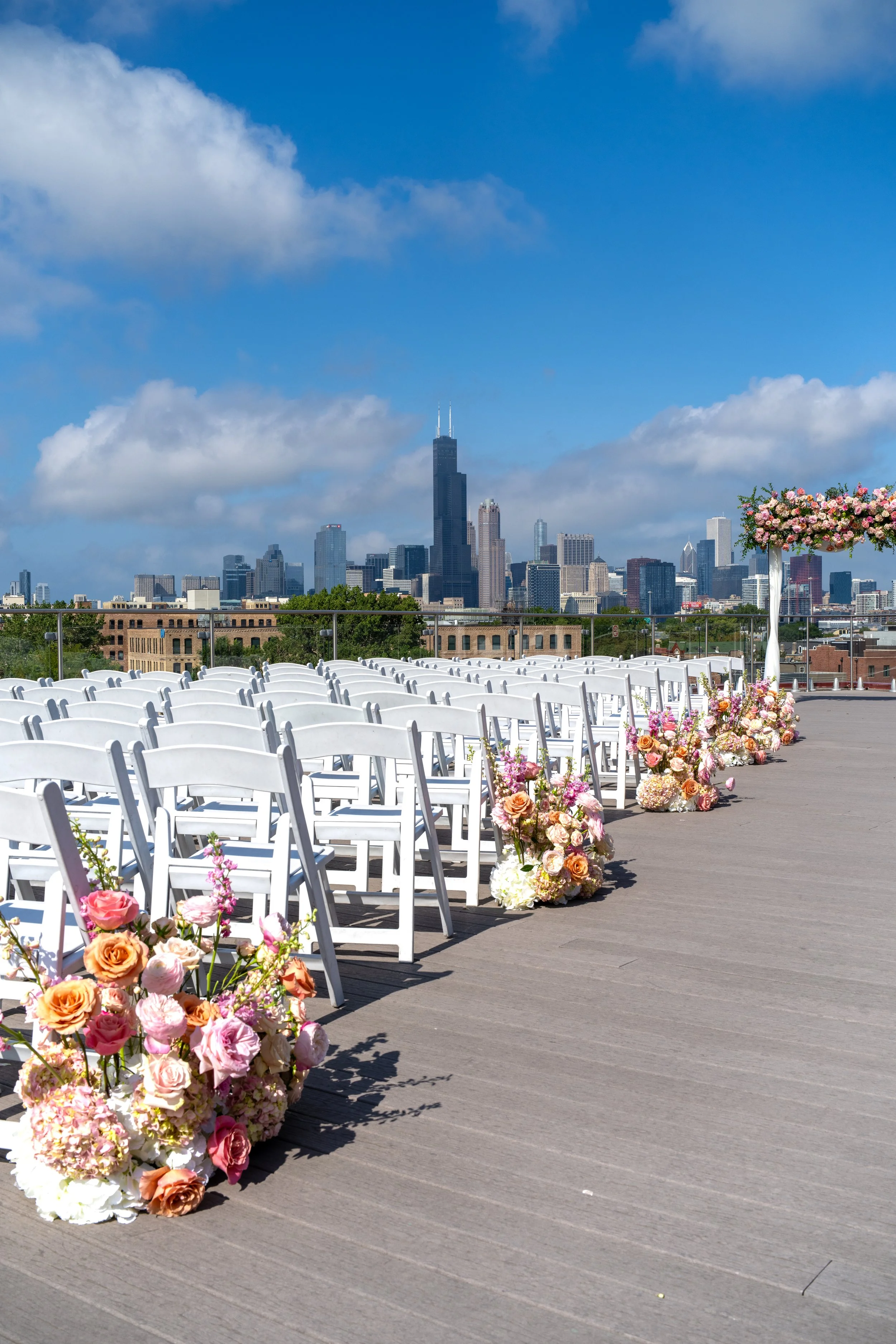 Outdoor wedding ceremony setup on a rooftop with white chairs, floral arrangements, and the Chicago skyline in the background, including the Willis Tower.