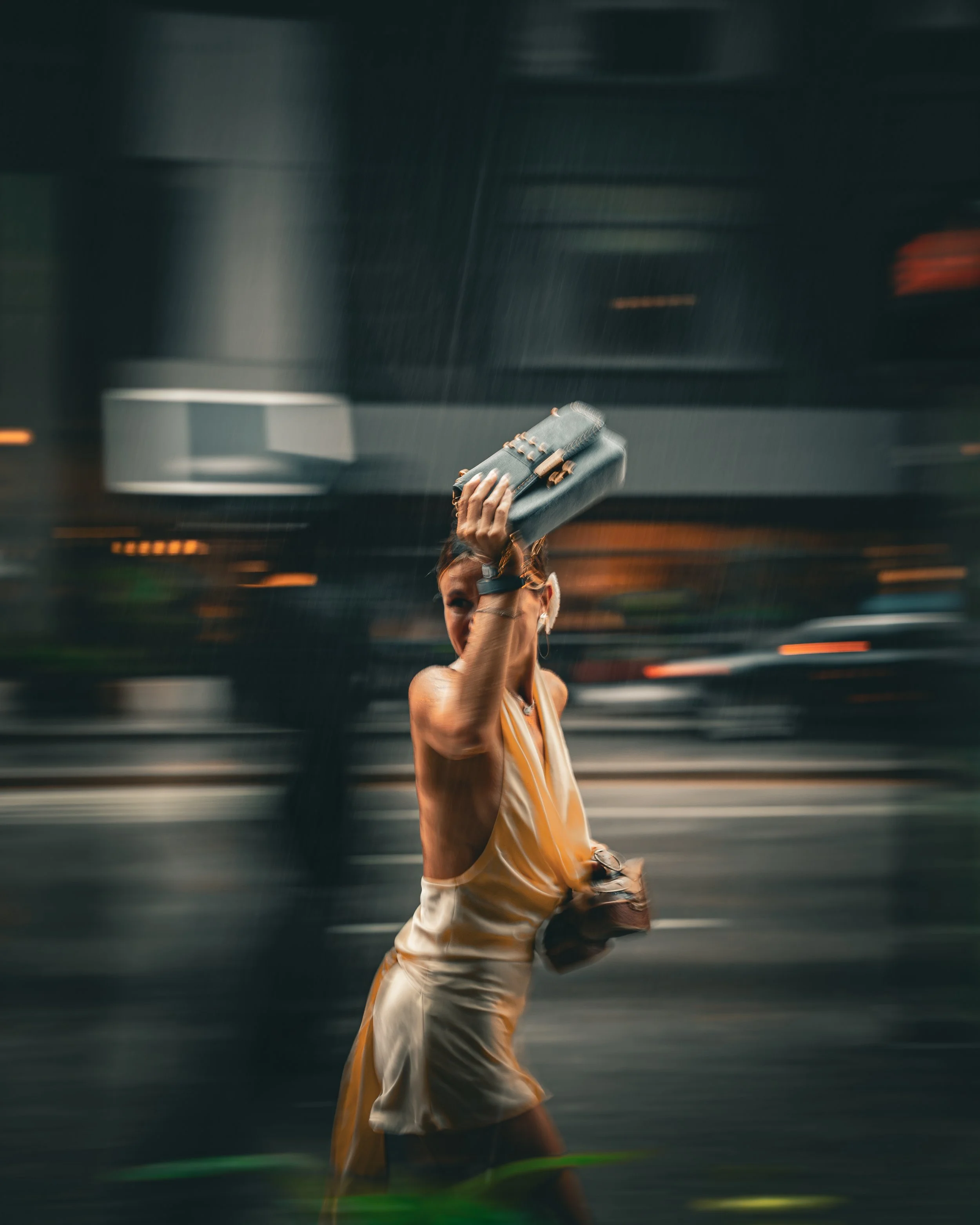 A woman is walking outdoors at night, holding a gray clutch bag above her head. She is wearing a beige dress with spaghetti straps and appears to be smiling. The background is blurred with moving lights and dark buildings.