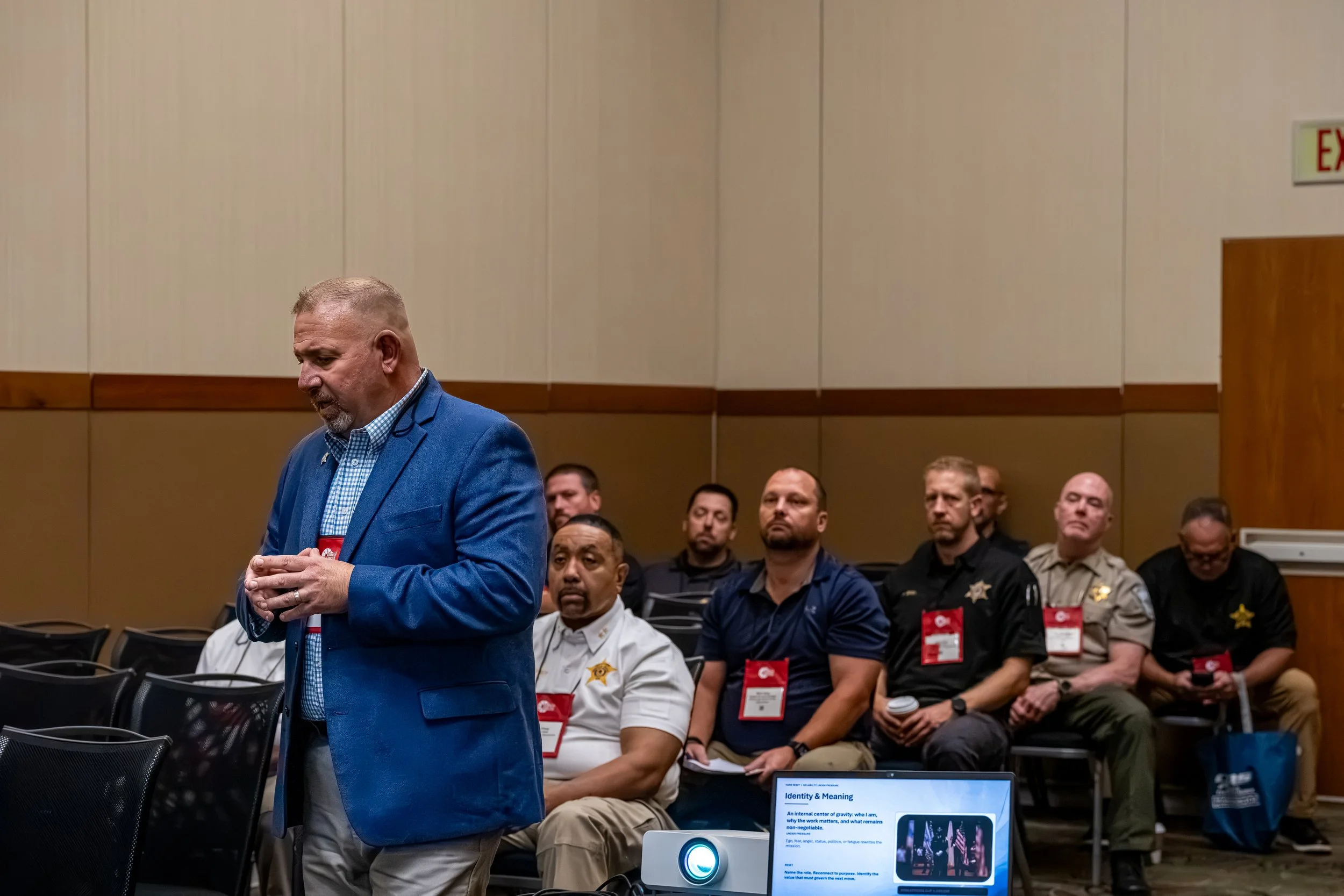A man in a blue suit is standing and speaking at a conference or seminar, with several seated attendees behind him, including law enforcement officers and civilians, in a conference room with beige walls.