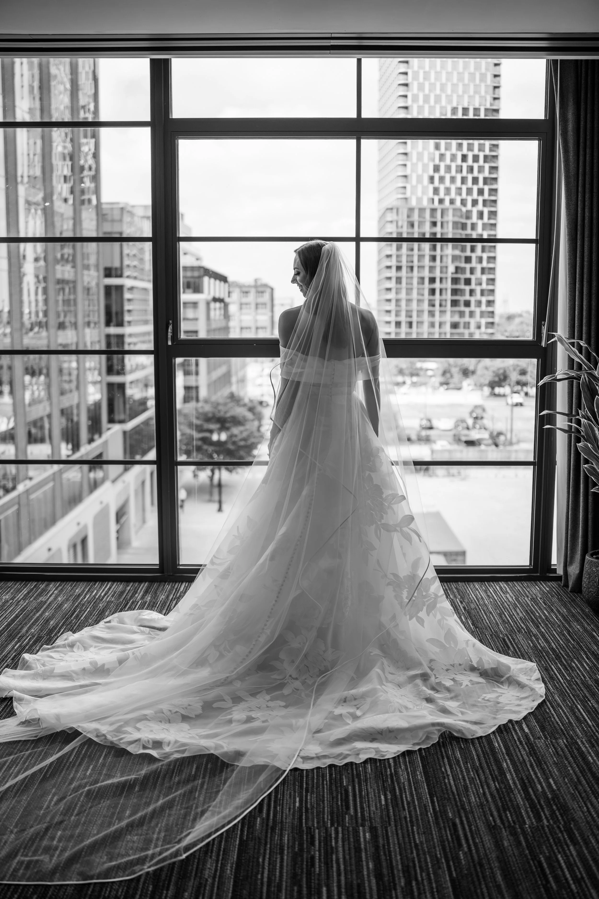 Black and white photo of a bride in a wedding dress with a long train and veil, standing by a large window in an urban setting.
