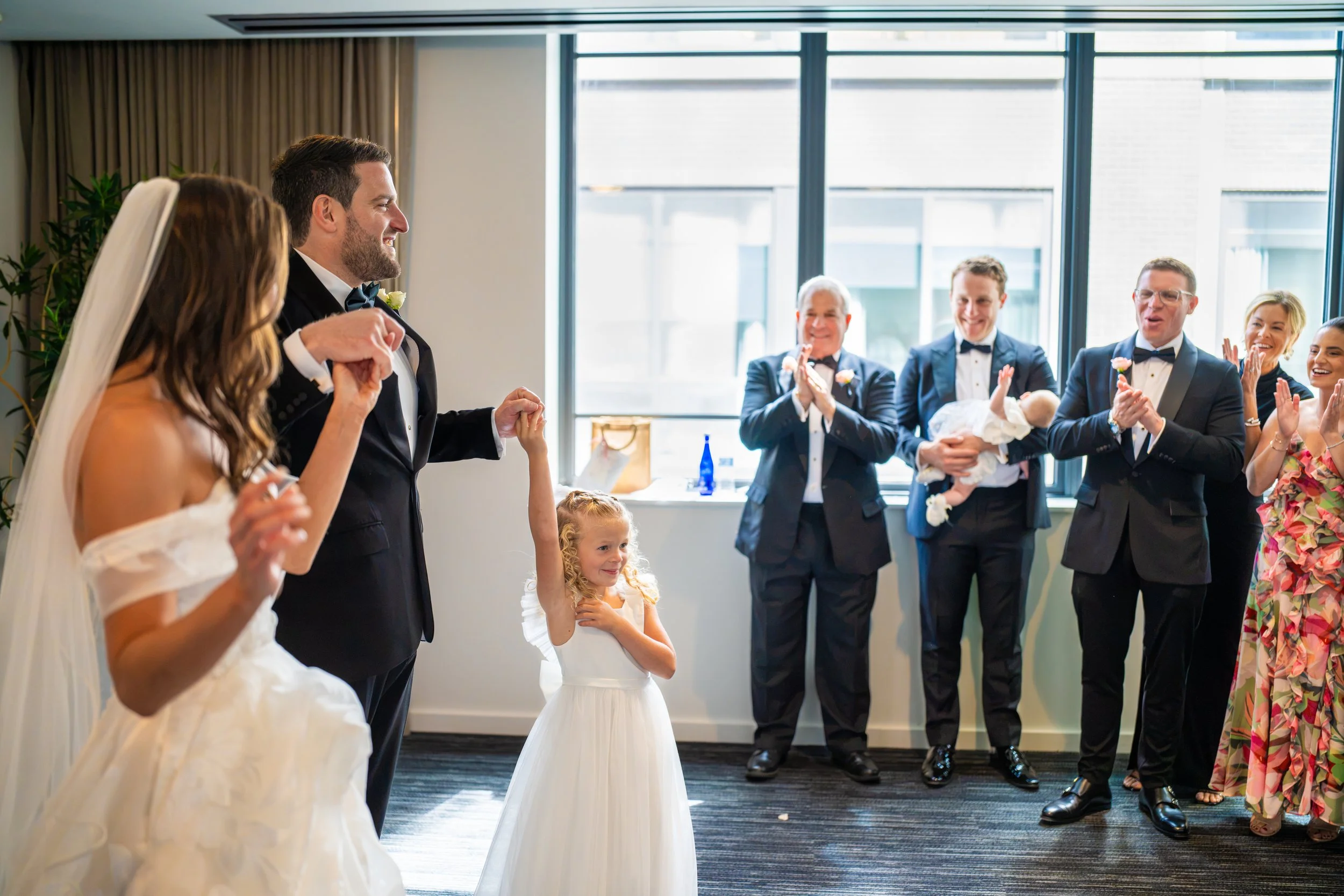 A group of wedding guests and children celebrating indoors near large windows with city views. The bride and groom are in the foreground, smiling.