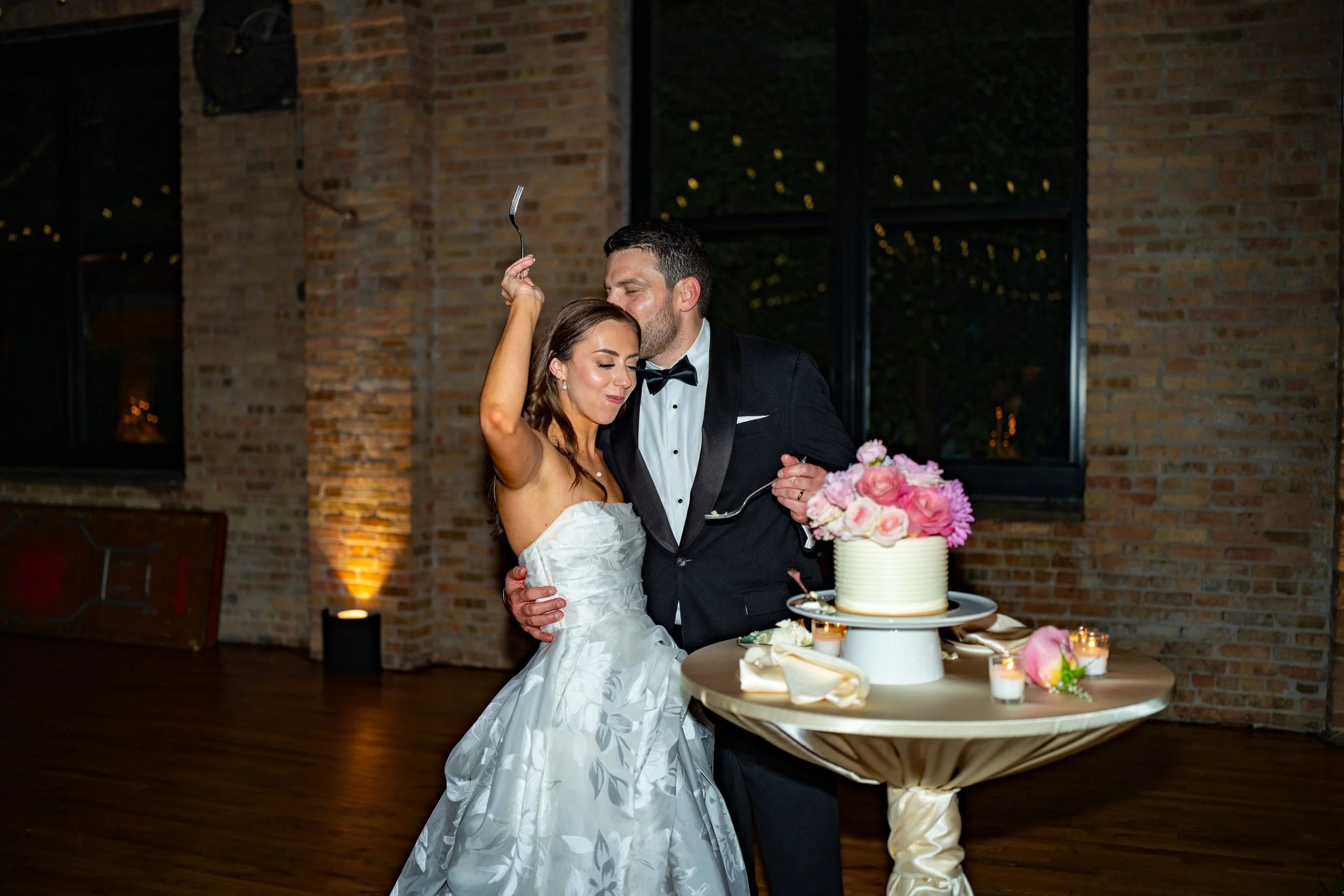 A bride and groom are dancing together at a wedding reception, with a wedding cake and pink floral arrangement on a table in front of them. They are in an indoor venue with brick walls and large windows.