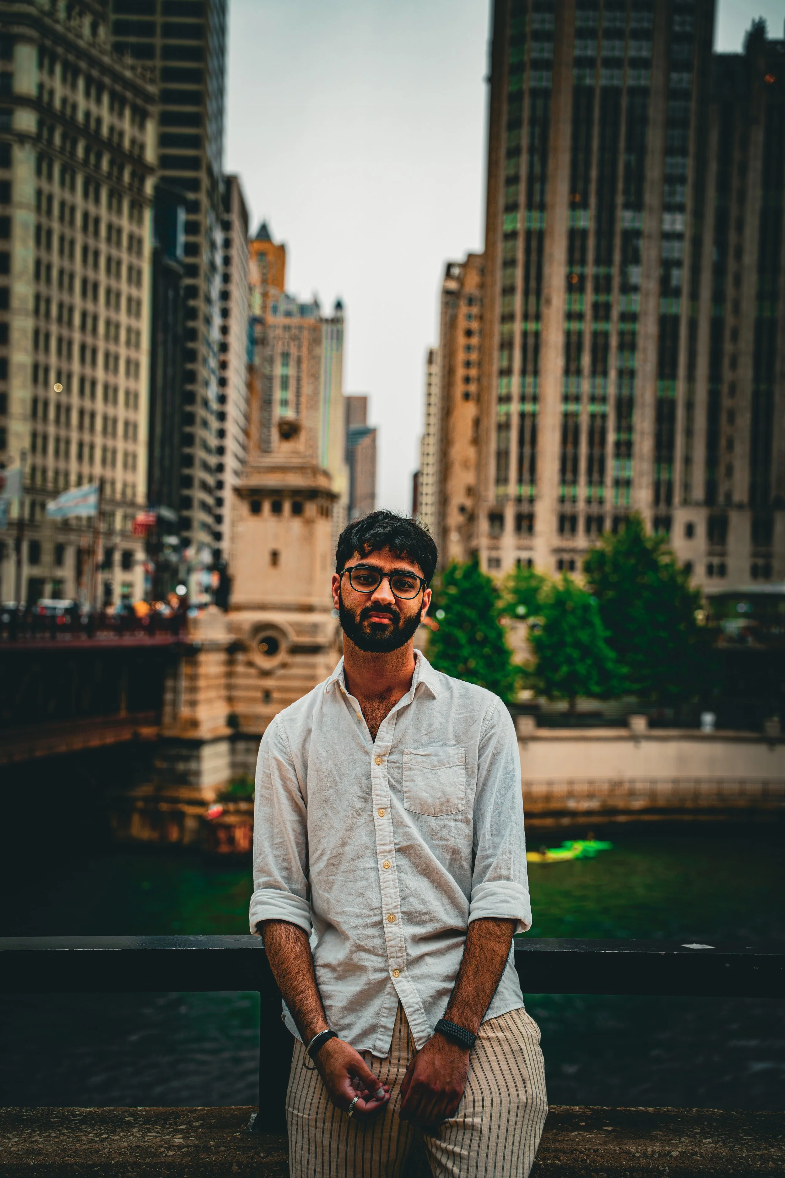 A man with glasses and a beard standing on a bridge with tall city buildings in the background during daytime.