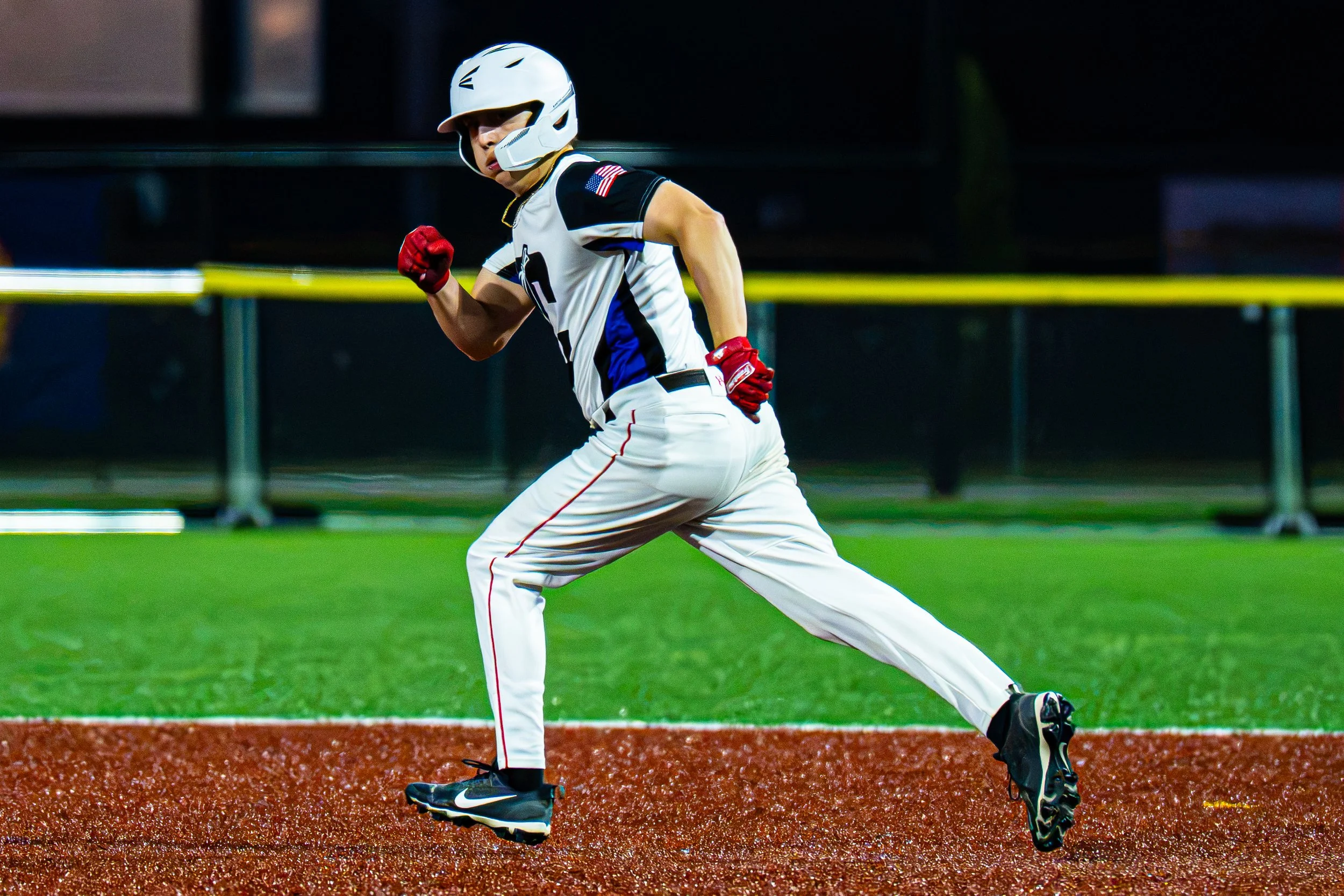 A baseball player running on the field, wearing a white uniform with black and blue accents, a white helmet, and red gloves, at night under stadium lights.