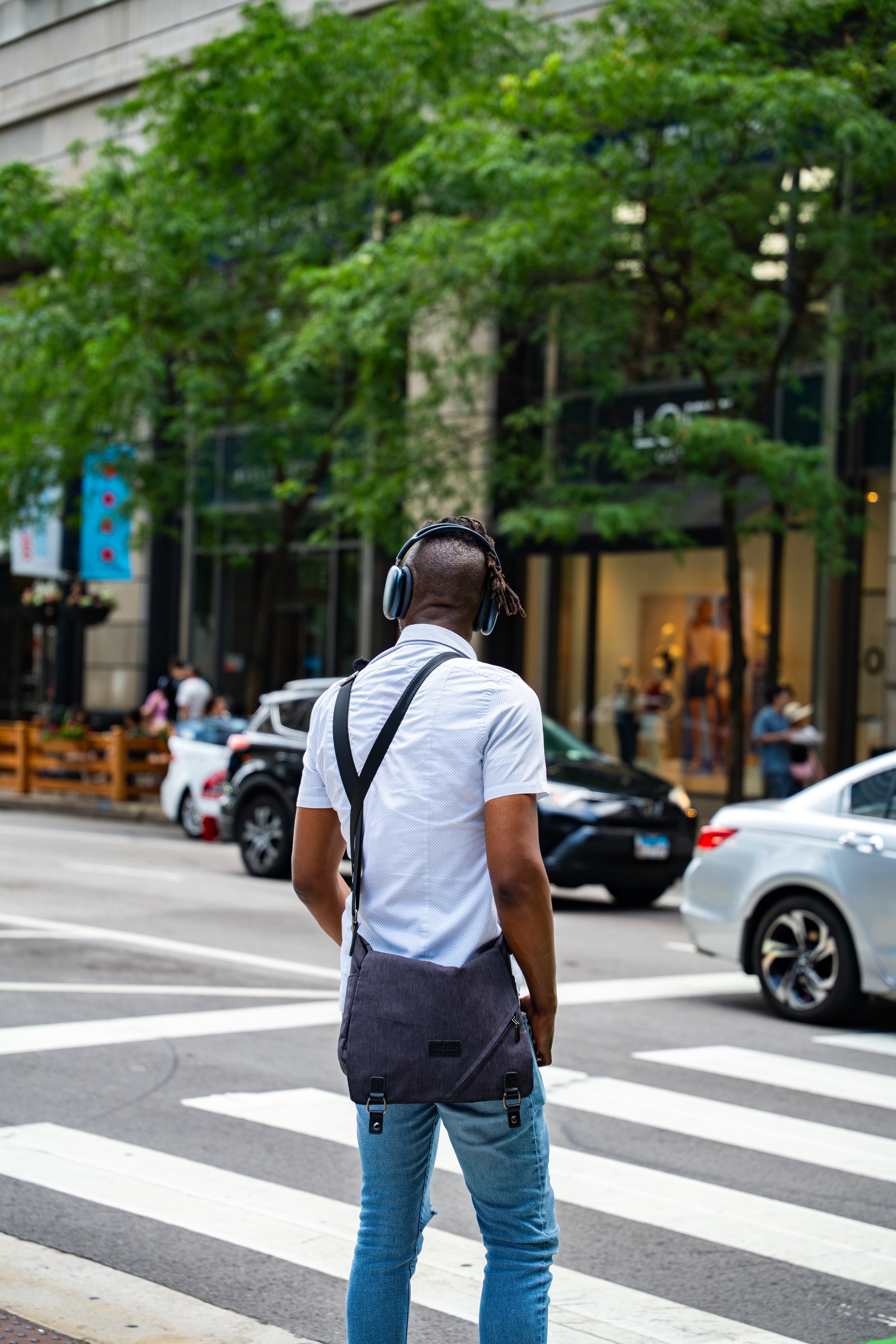 A man with dreadlocks wearing a white shirt, blue jeans, and a black backpack crossing a street with cars and pedestrians around, while listening to music with headphones, in an urban setting with buildings and trees.