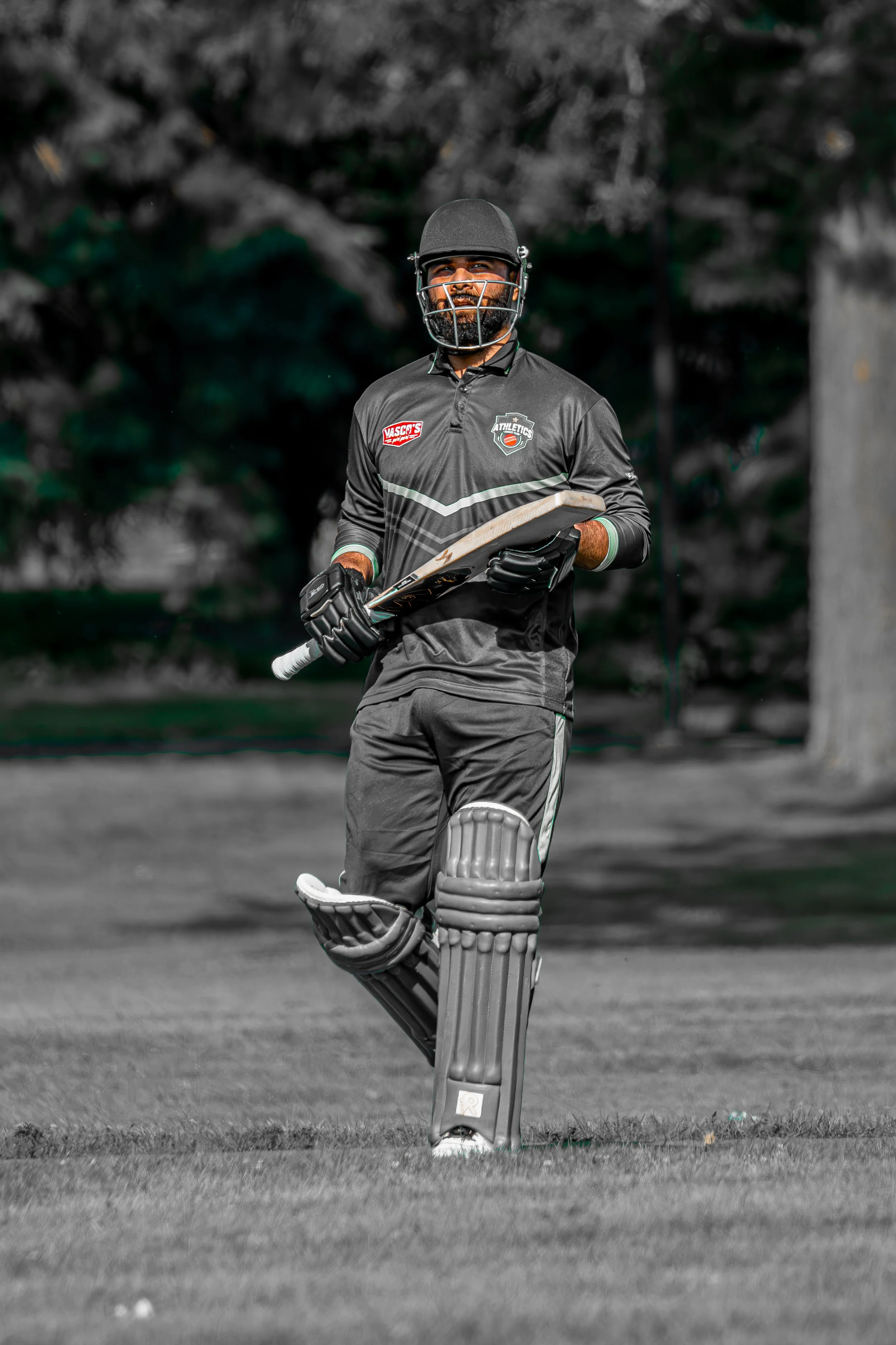 A male cricket player dressed in black practice gear, holding a cricket bat, with a helmet and protective pads, standing on a cricket field.