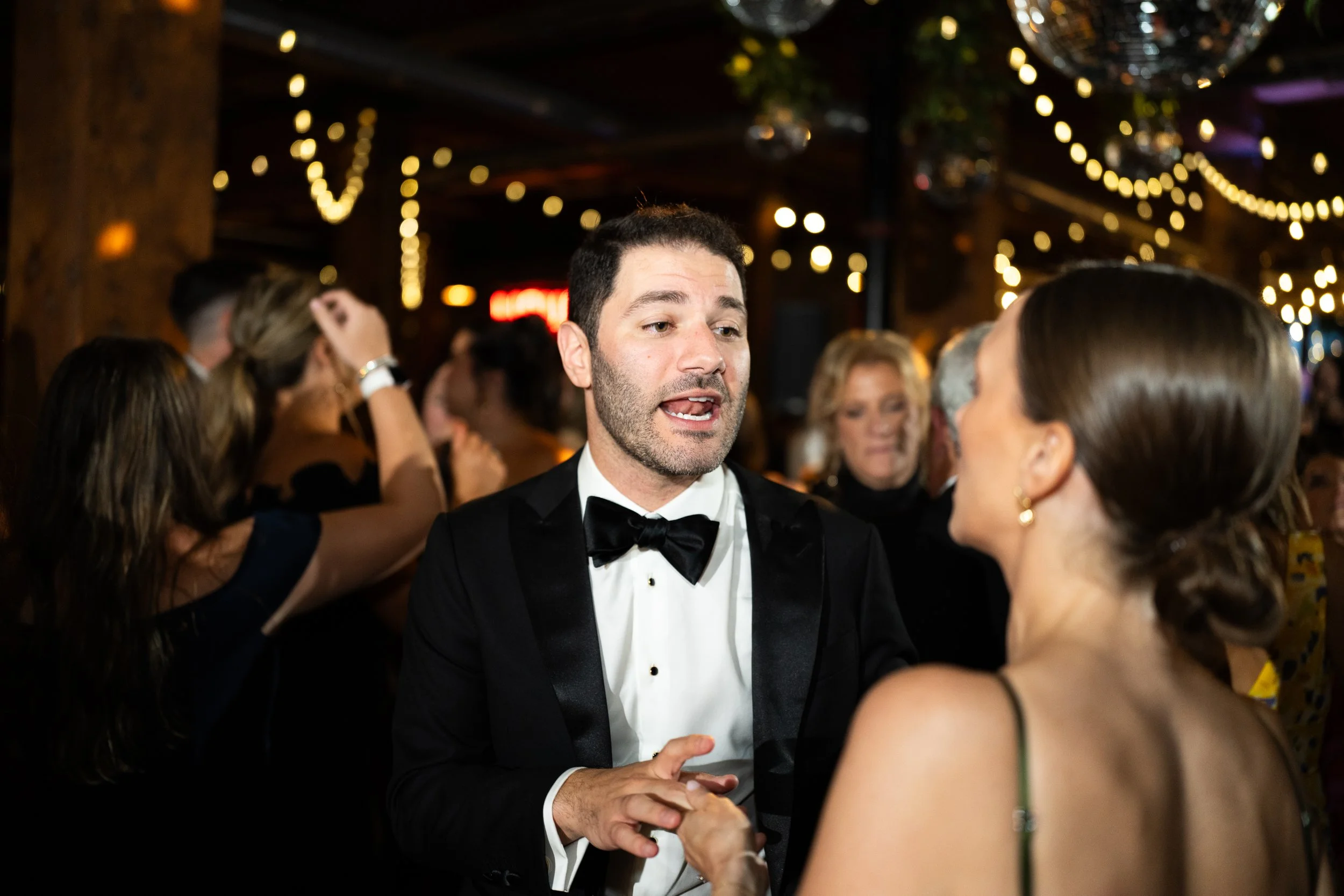 A man in a tuxedo and bowtie dancing and talking to a woman at a crowded party or celebration, with string lights and disco balls hanging from the ceiling in the background.