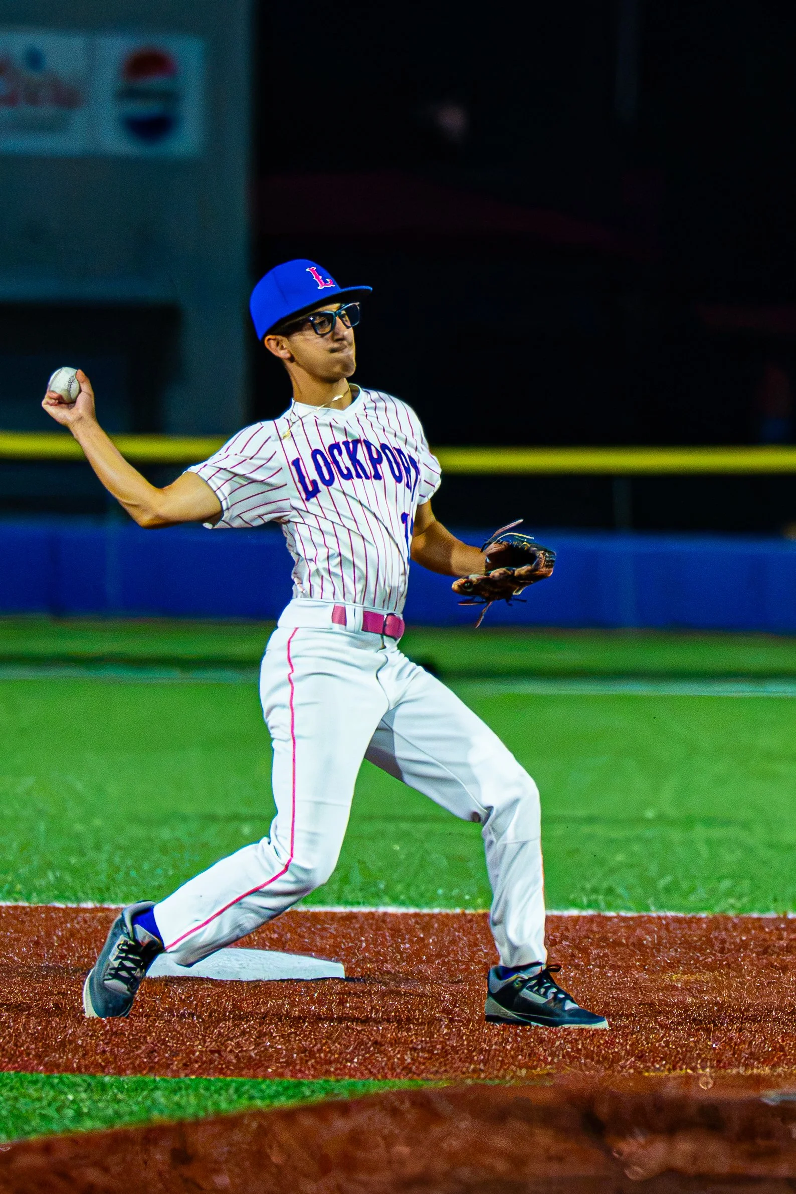 A baseball player in a striped uniform with 'LOCKPORT' written on it, wearing a blue cap with a red 'L', glasses, and black cleats, is preparing to throw a baseball from a base on a baseball field at night.