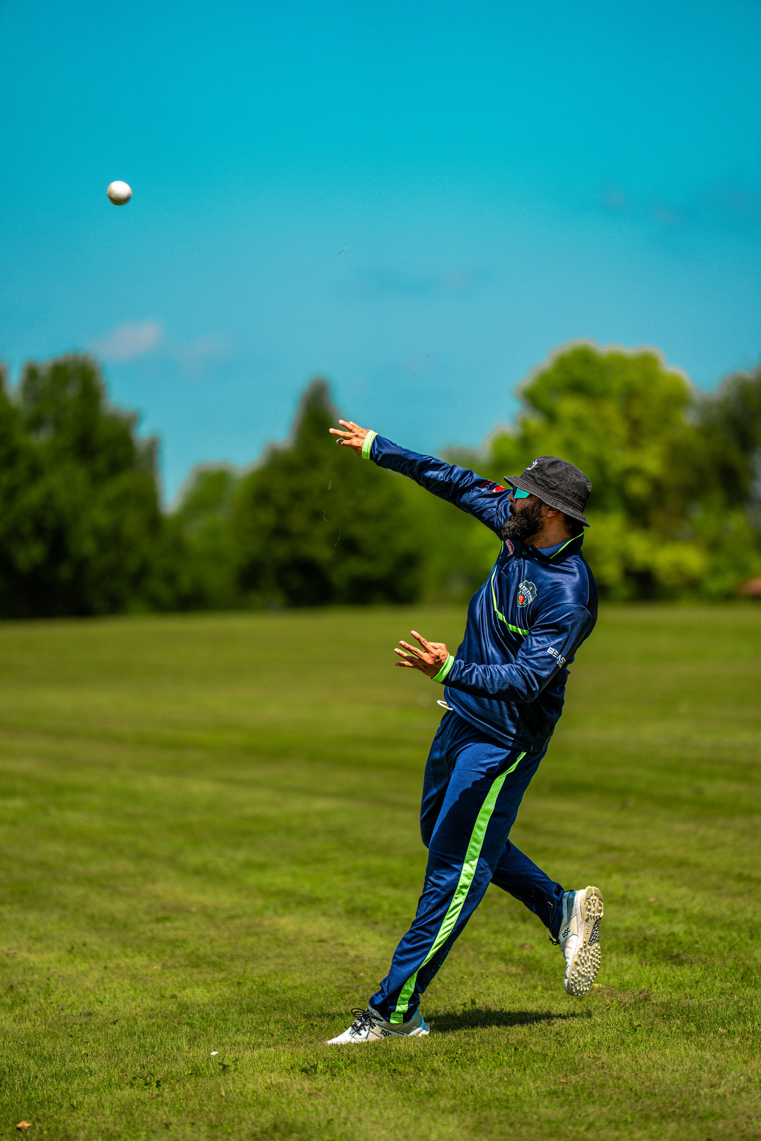 A man in a blue sports outfit and hat playing cricket on a grassy field, preparing to catch a flying ball in a clear outdoor setting with trees and blue sky.