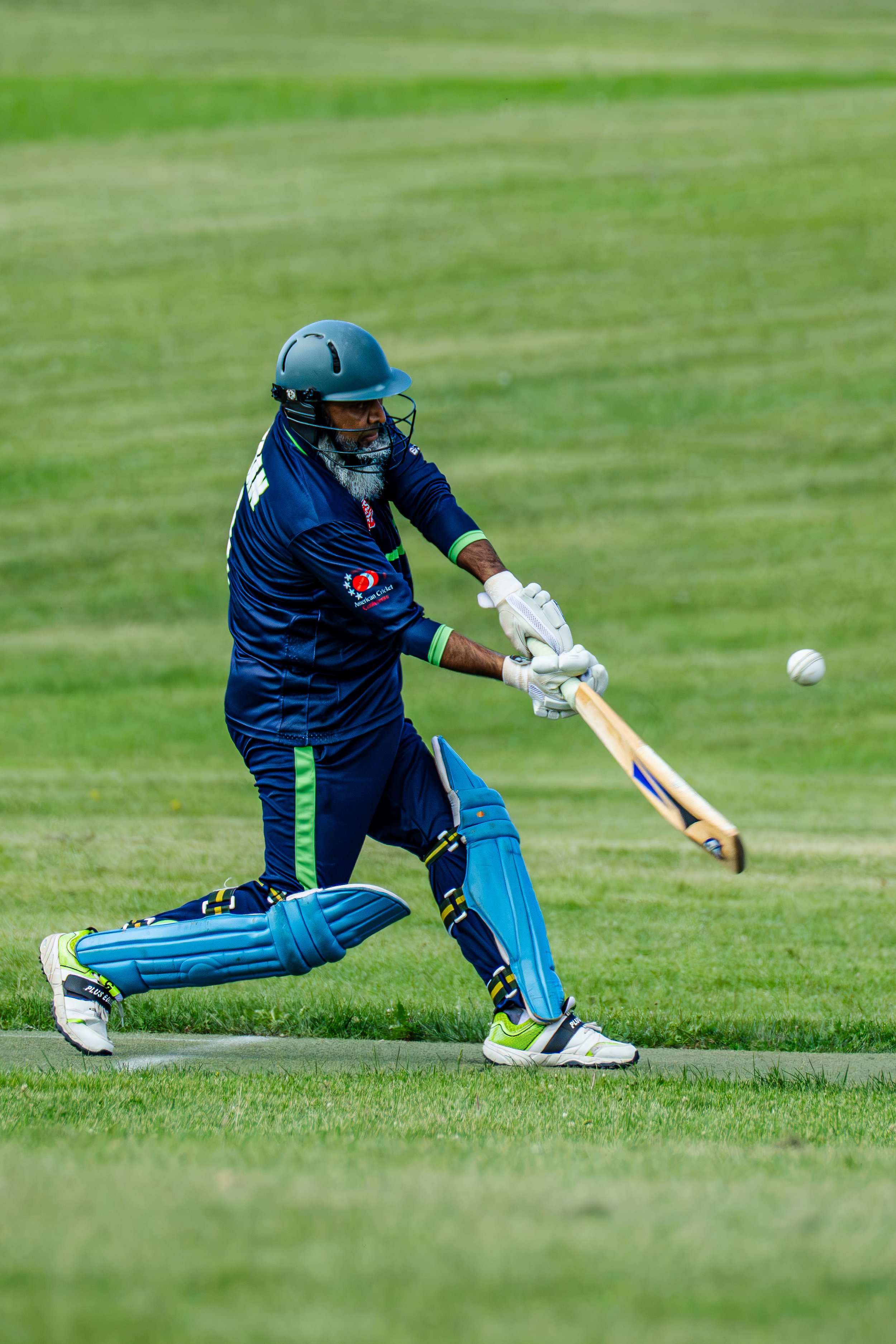 A man dressed in blue cricket uniform, wearing a helmet and gloves, is in motion batting in a cricket game on a grassy field.