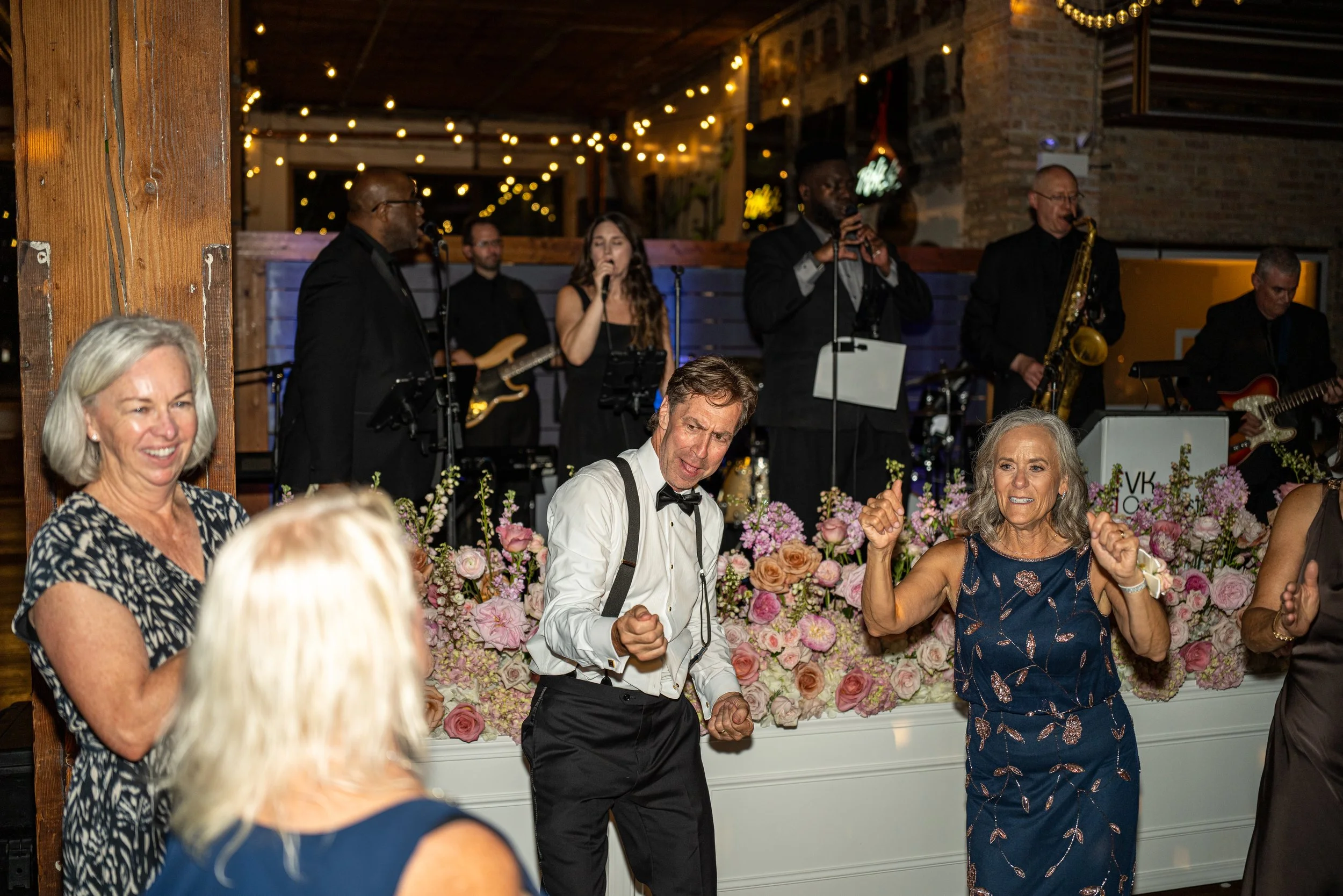 People dancing and enjoying themselves at a wedding reception with a live band performing in the background and floral decorations on a table.