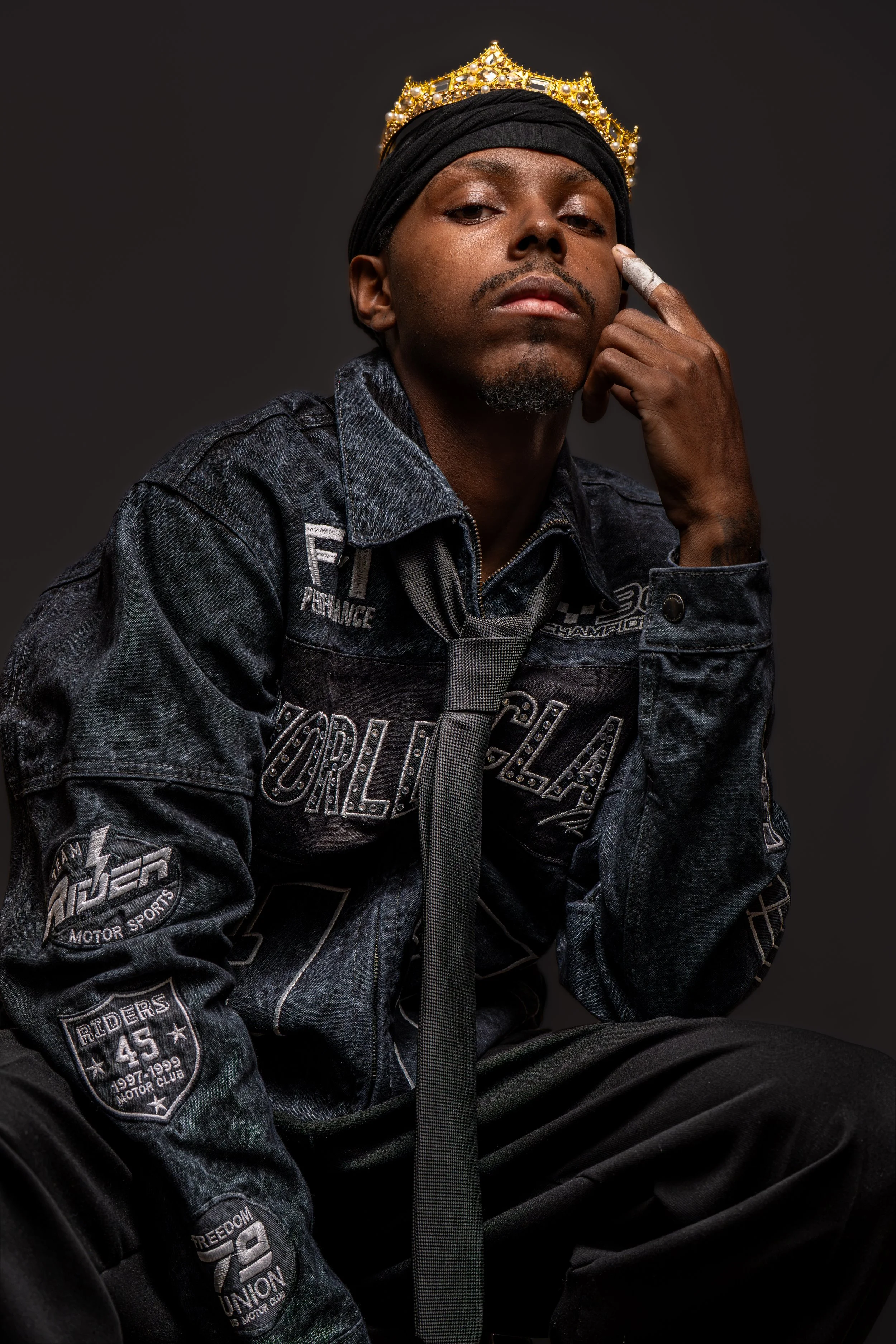 A man with dark skin wearing a black hoodie, a graphic denim jacket, and a black headscarf. He has a gold crown-style headpiece decorated with pearls on his head. He is sitting and looking directly at the camera with a serious expression, with his right hand touching his face, showing a ring on his finger.