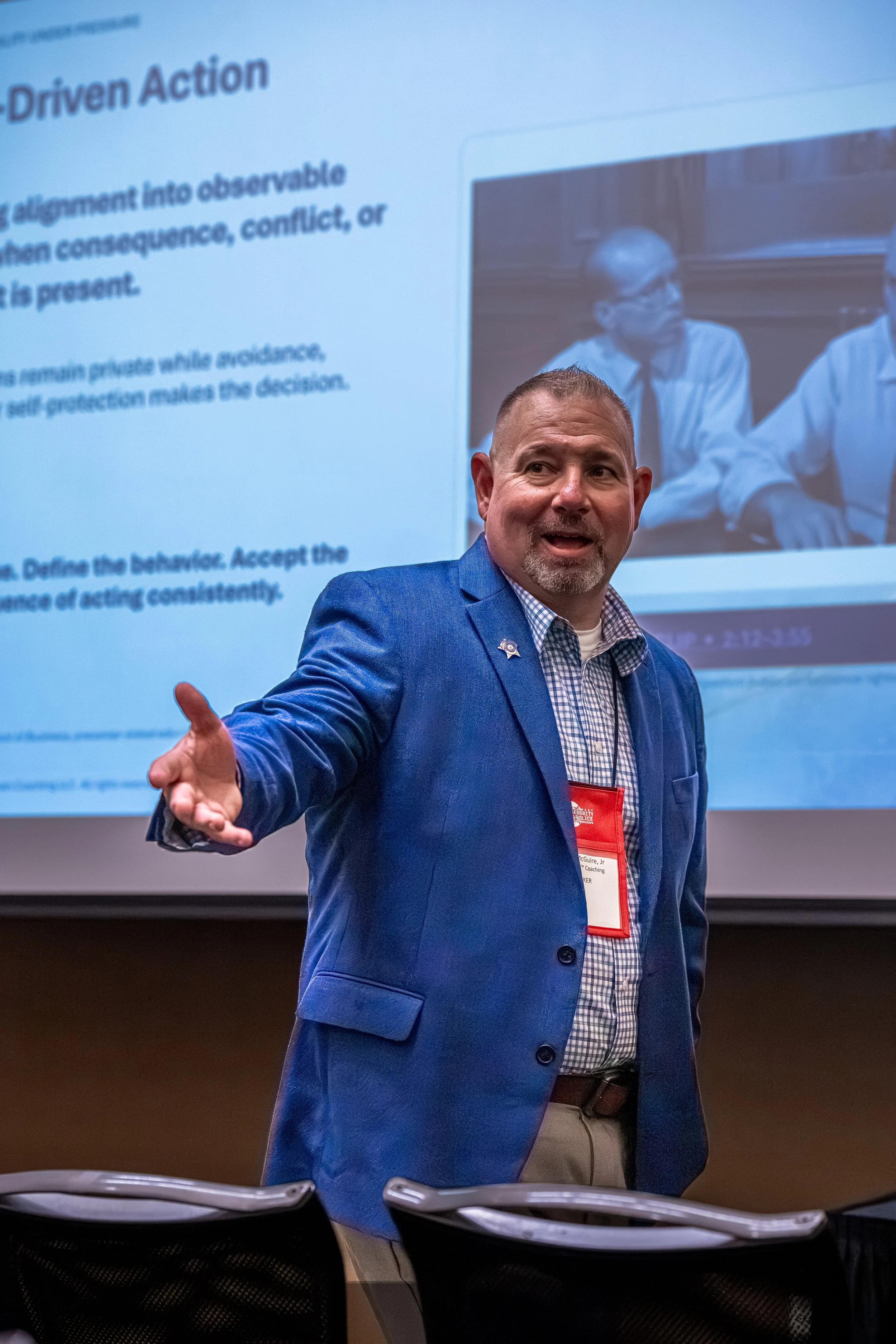 A man in a blue blazer and checkered shirt speaking in front of a presentation slide with text and a photo of two people in white shirts. The man has short hair and is gesturing with his right hand.