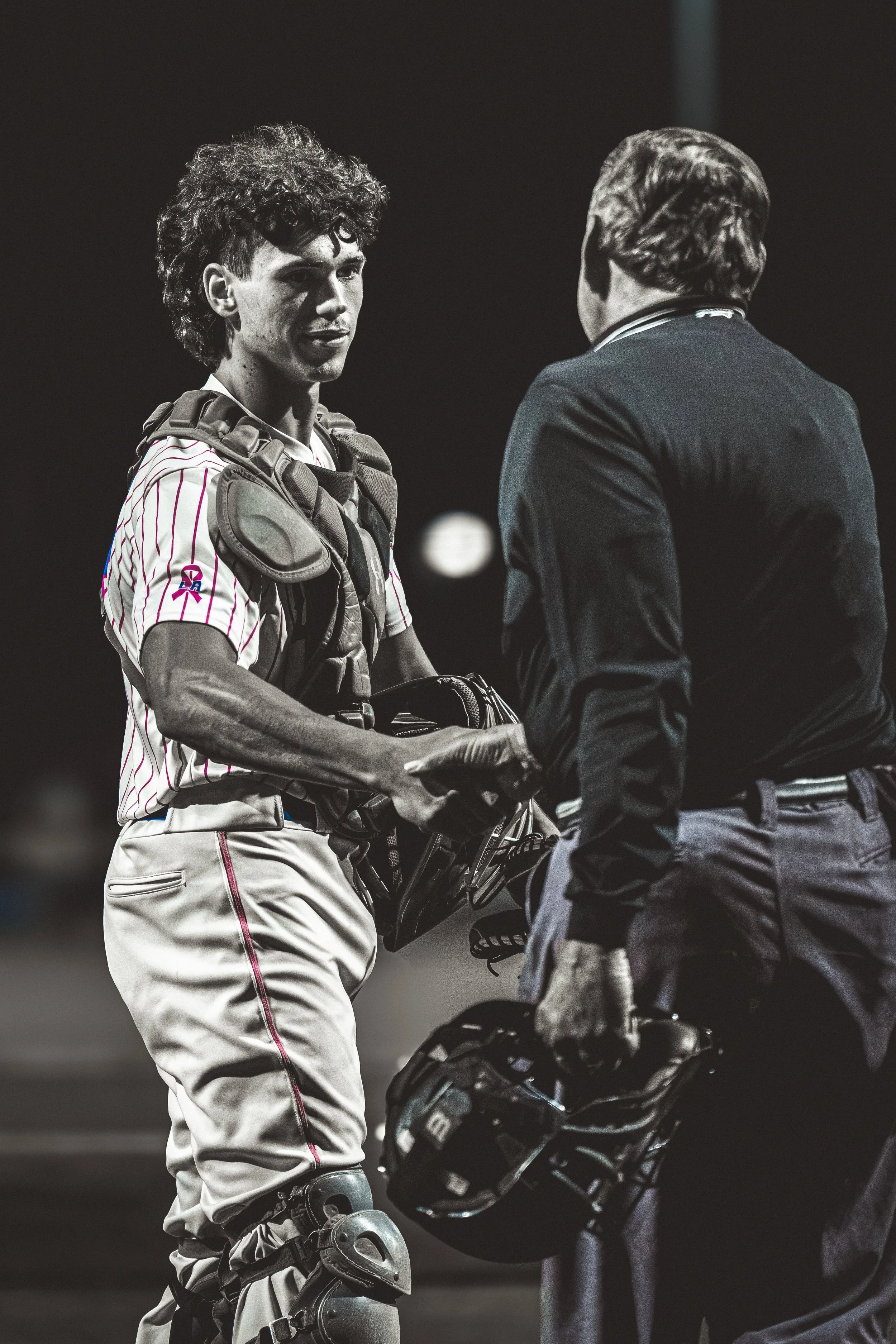 A young baseball player in uniform with catcher's gear, shaking hands with a man in formal attire, likely a coach or official, in a dark setting.