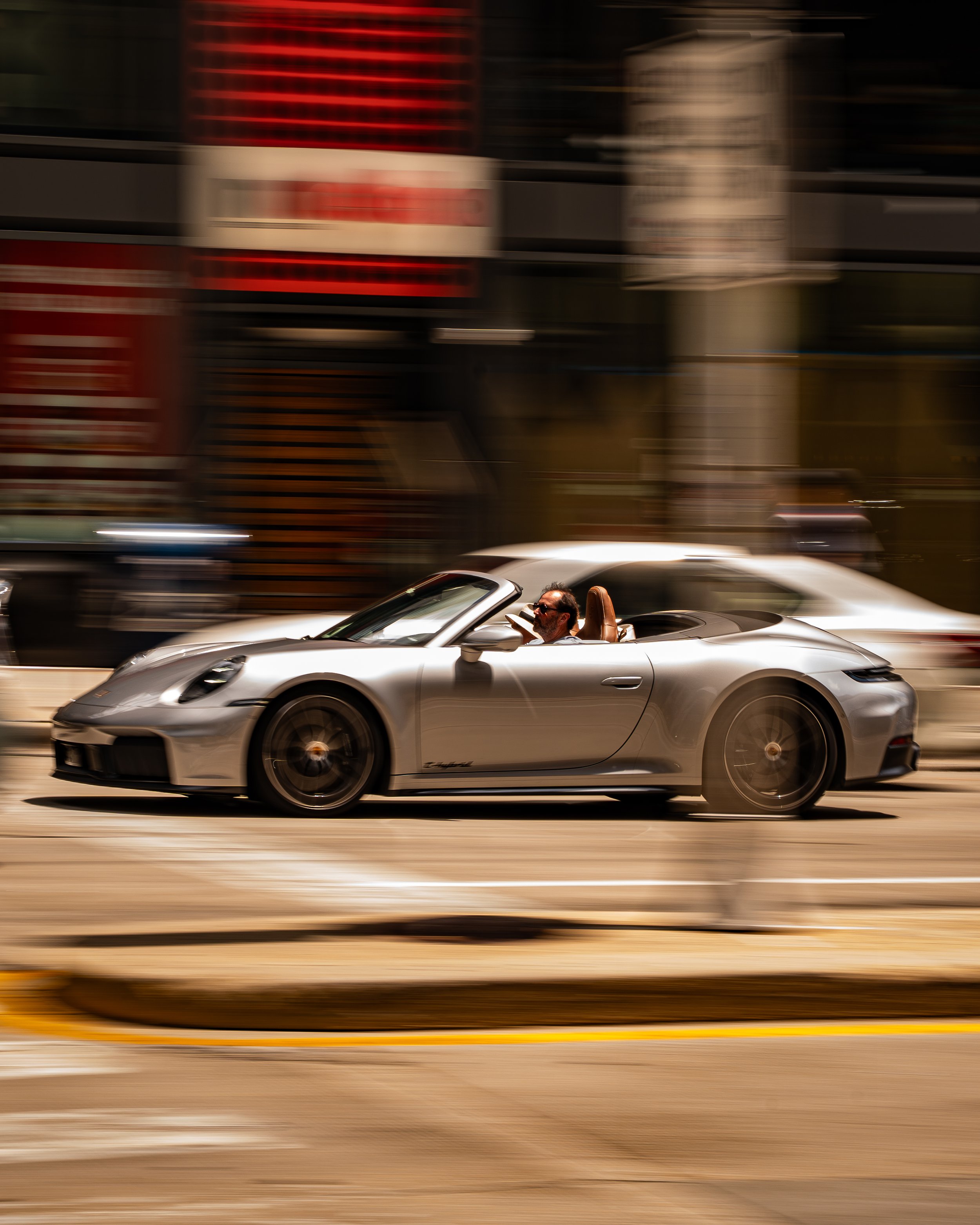 A man driving a silver convertible car on a city street, with blurred buildings and traffic in the background.