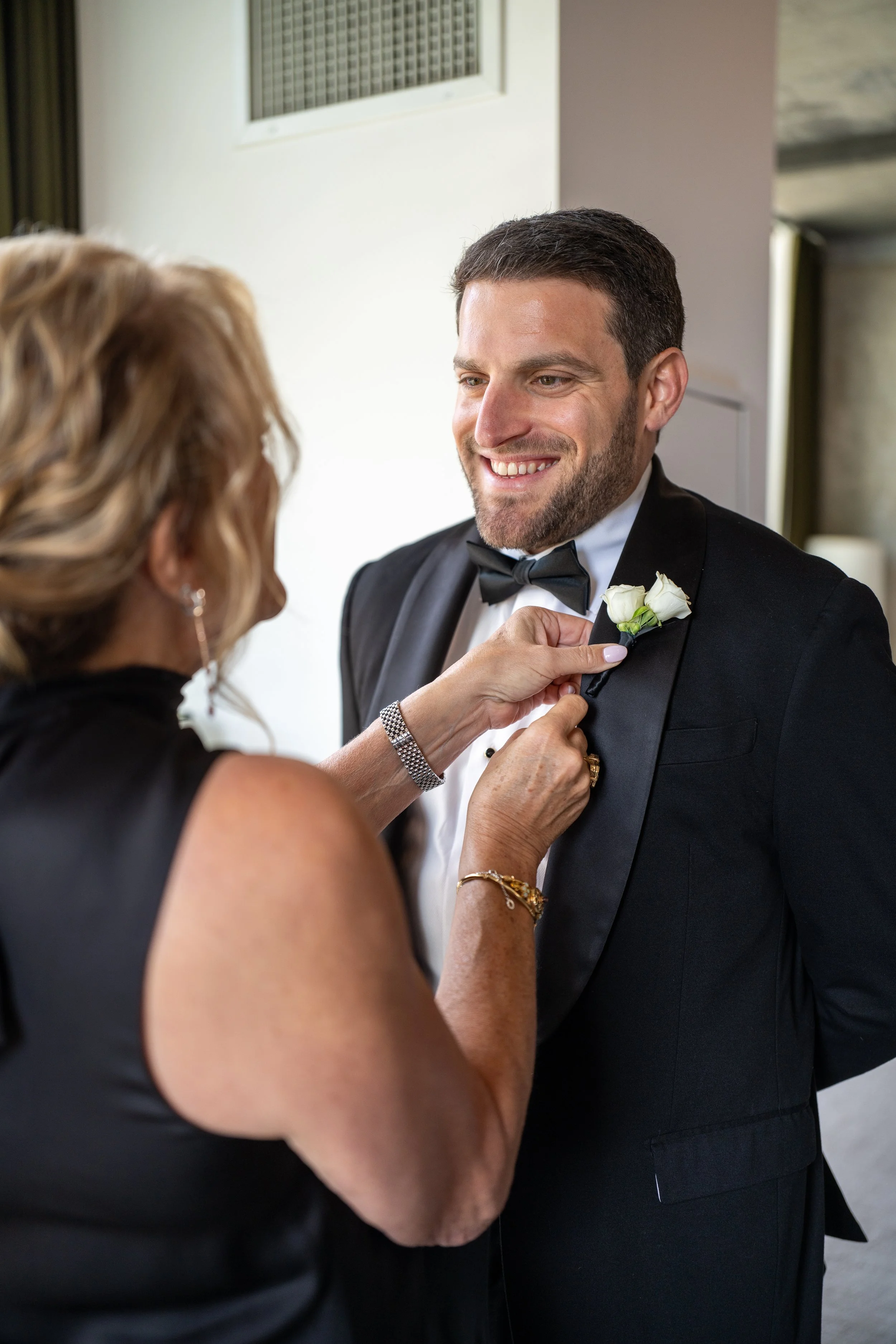 A man in a tuxedo smiling as a woman pins a white boutonniere onto his lapel.