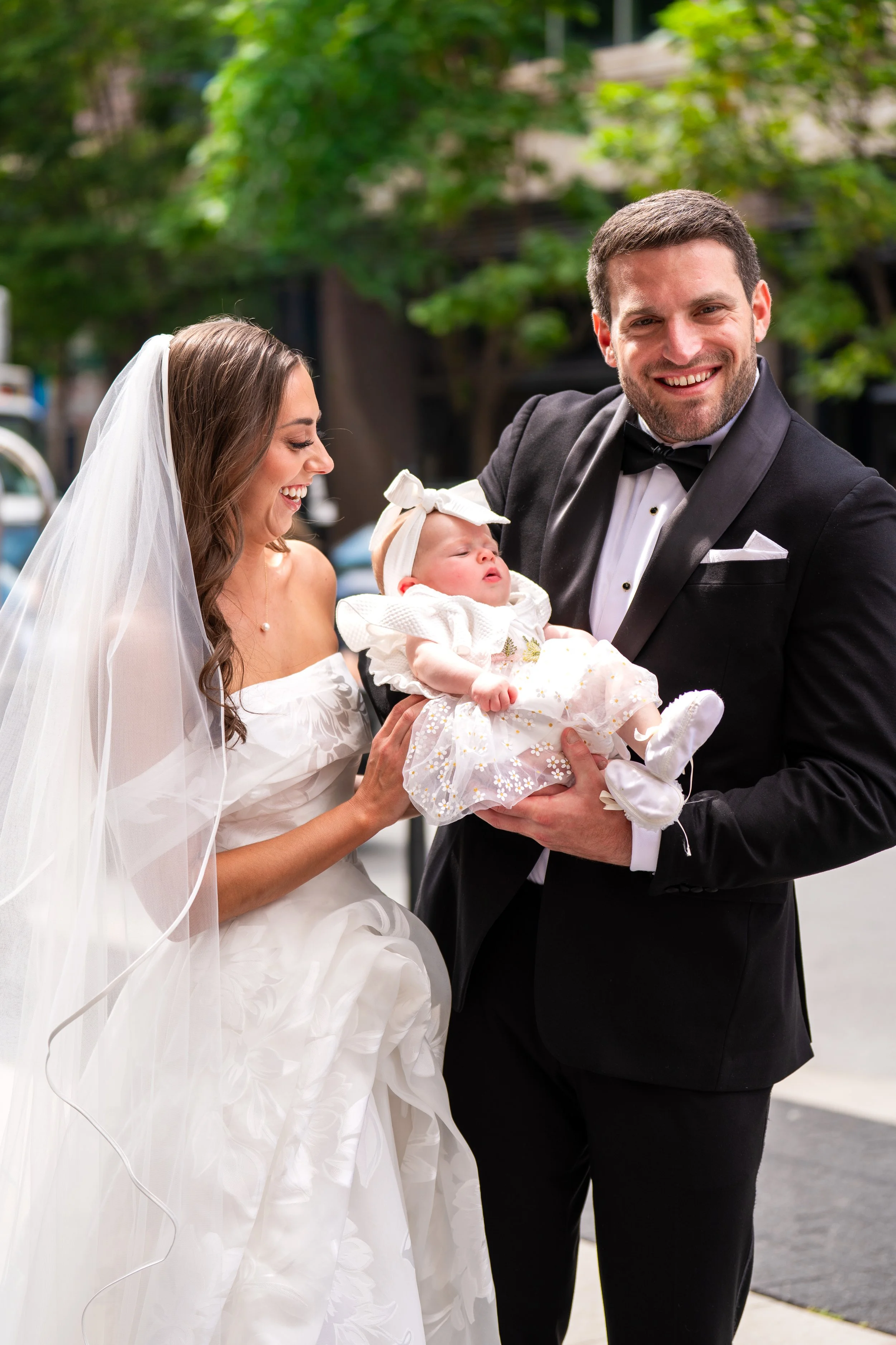 A smiling man in a tuxedo holding a baby girl dressed in white, with a woman in a wedding dress and veil looking at the baby, outdoors with green trees in the background.
