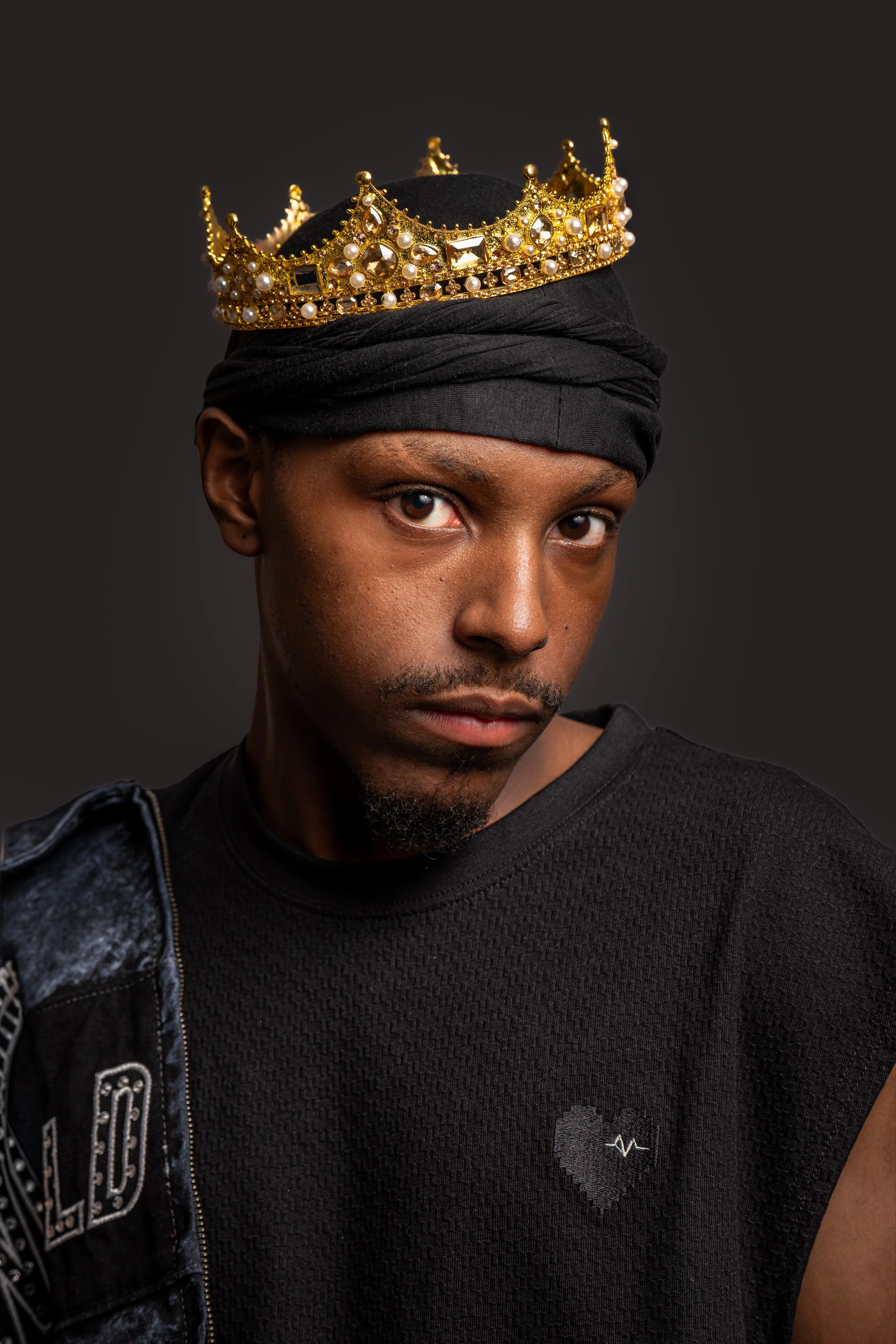 A person wearing a black head wrap and a black shirt with a small gray heart and heartbeat line graphic, with a gold crown adorned with pearls and gemstones on their head.