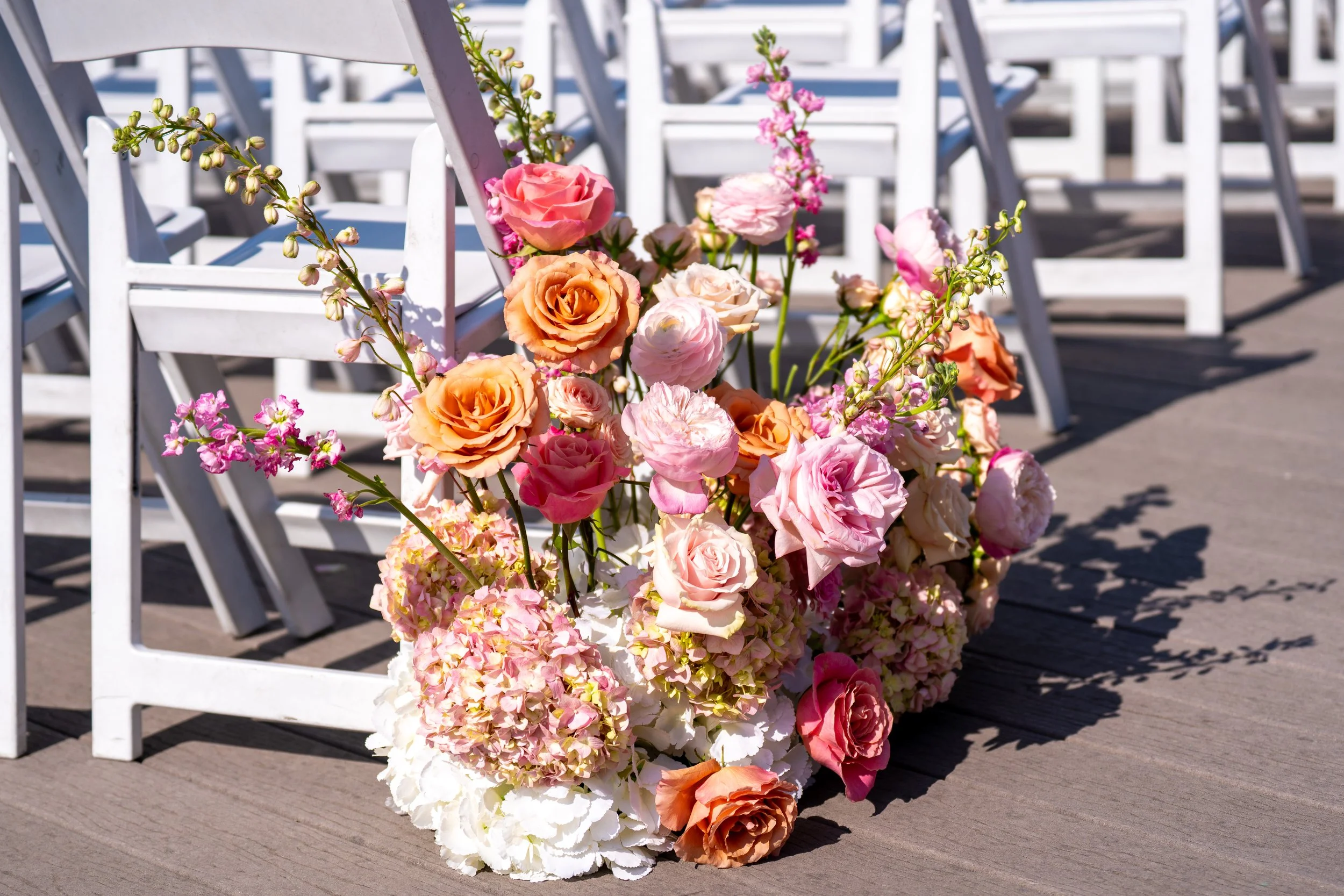 A large bouquet of various pink and peach roses and other flowers on a wooden deck, with white chairs in the background.