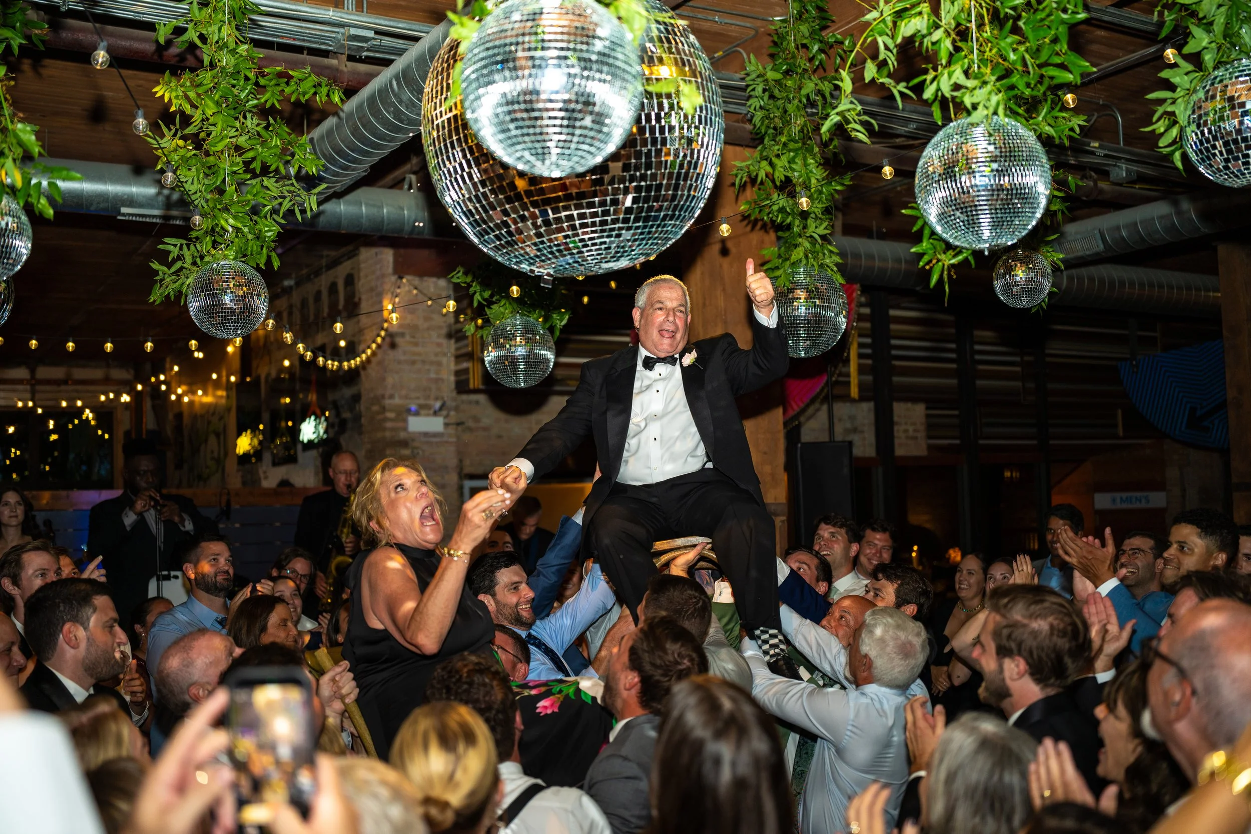 A lively celebration with a large crowd, featuring a man in a tuxedo and a woman in a black dress, both smiling. The man is sitting on someone's shoulders with one thumb up. There are disco balls and green plants hanging from the ceiling and string l